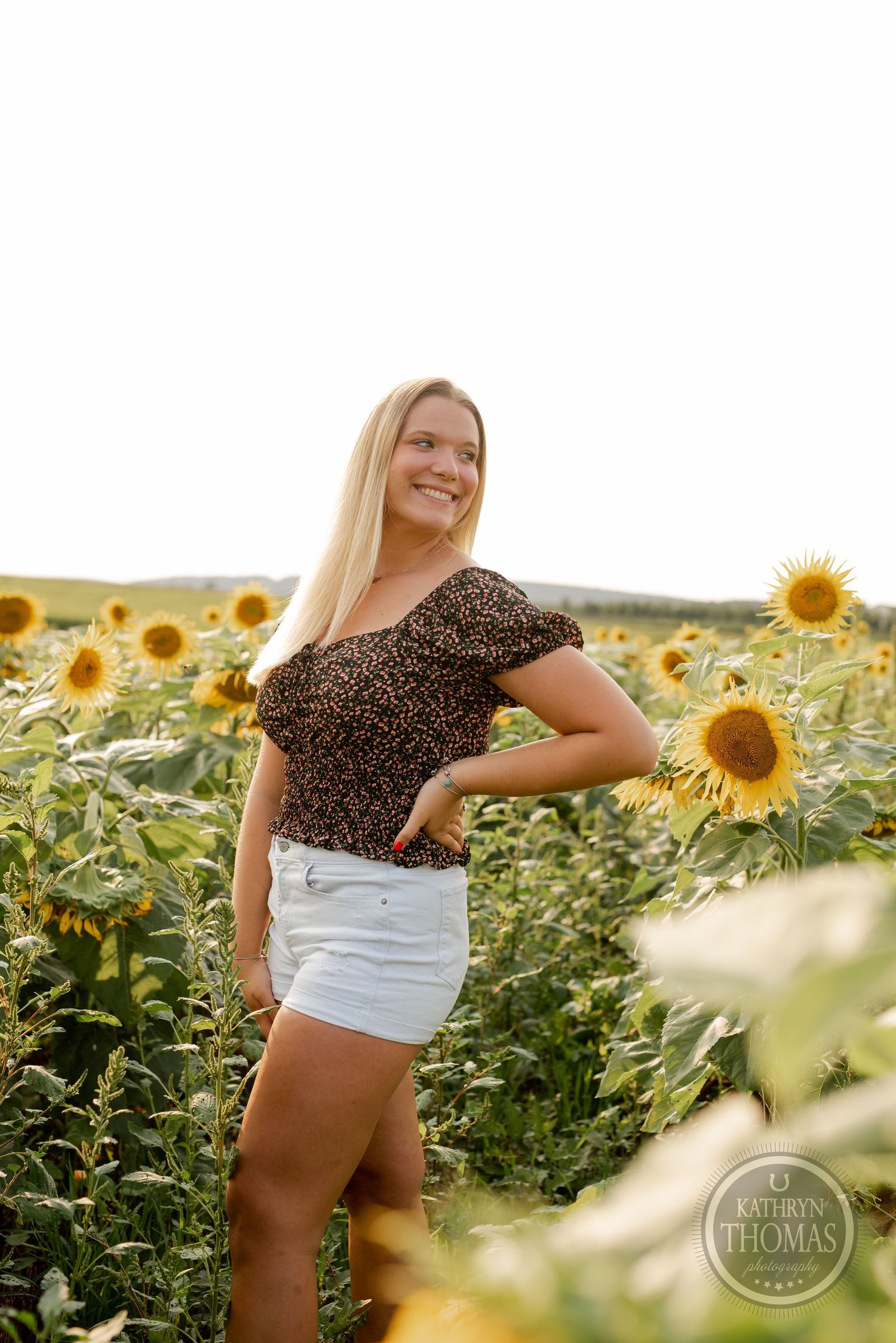 A person in a floral crop top and white shorts poses happily in a sunflower field at golden hour.