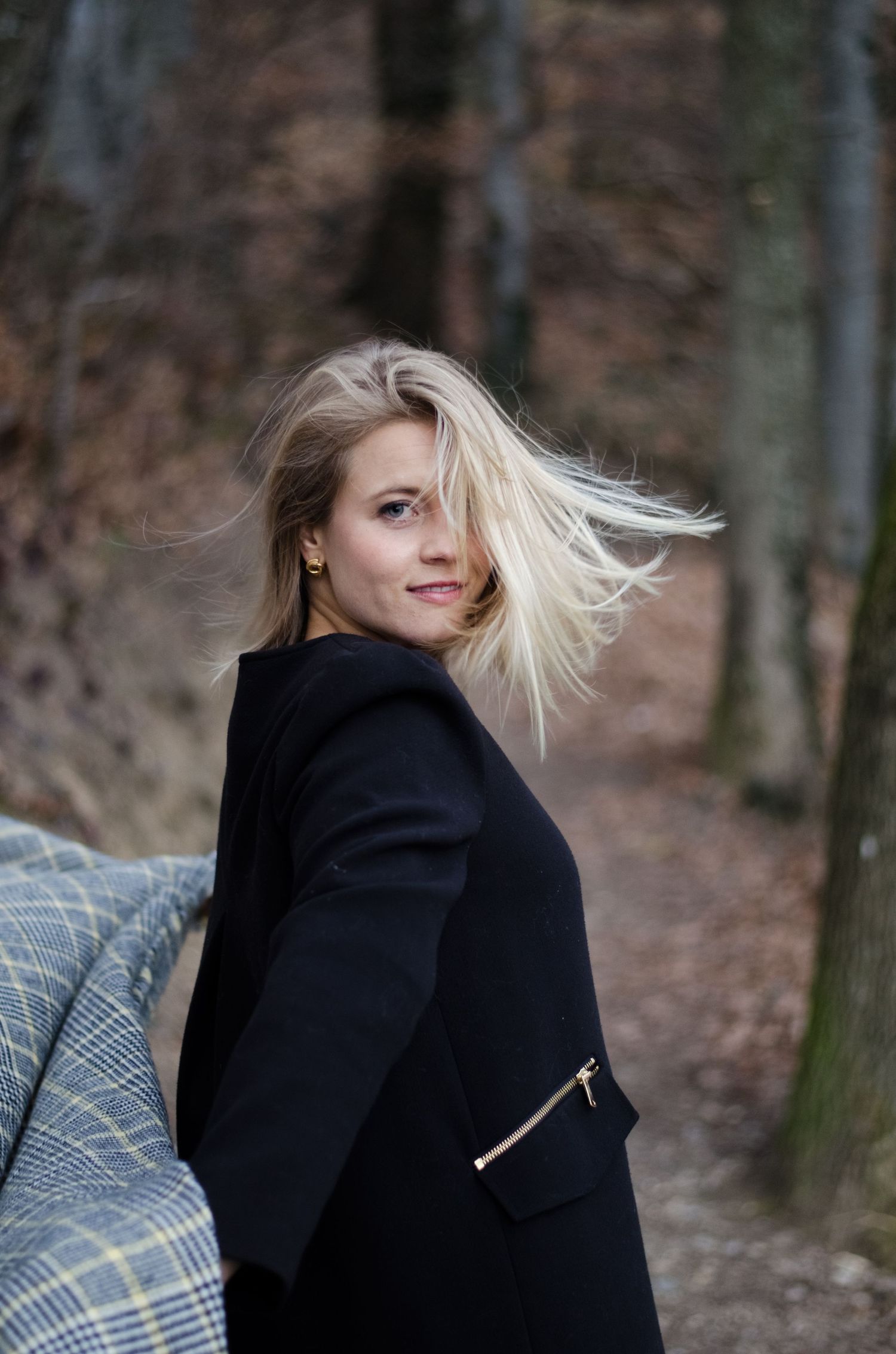 Person in a black coat sitting outdoors with blonde hair blowing in the wind.