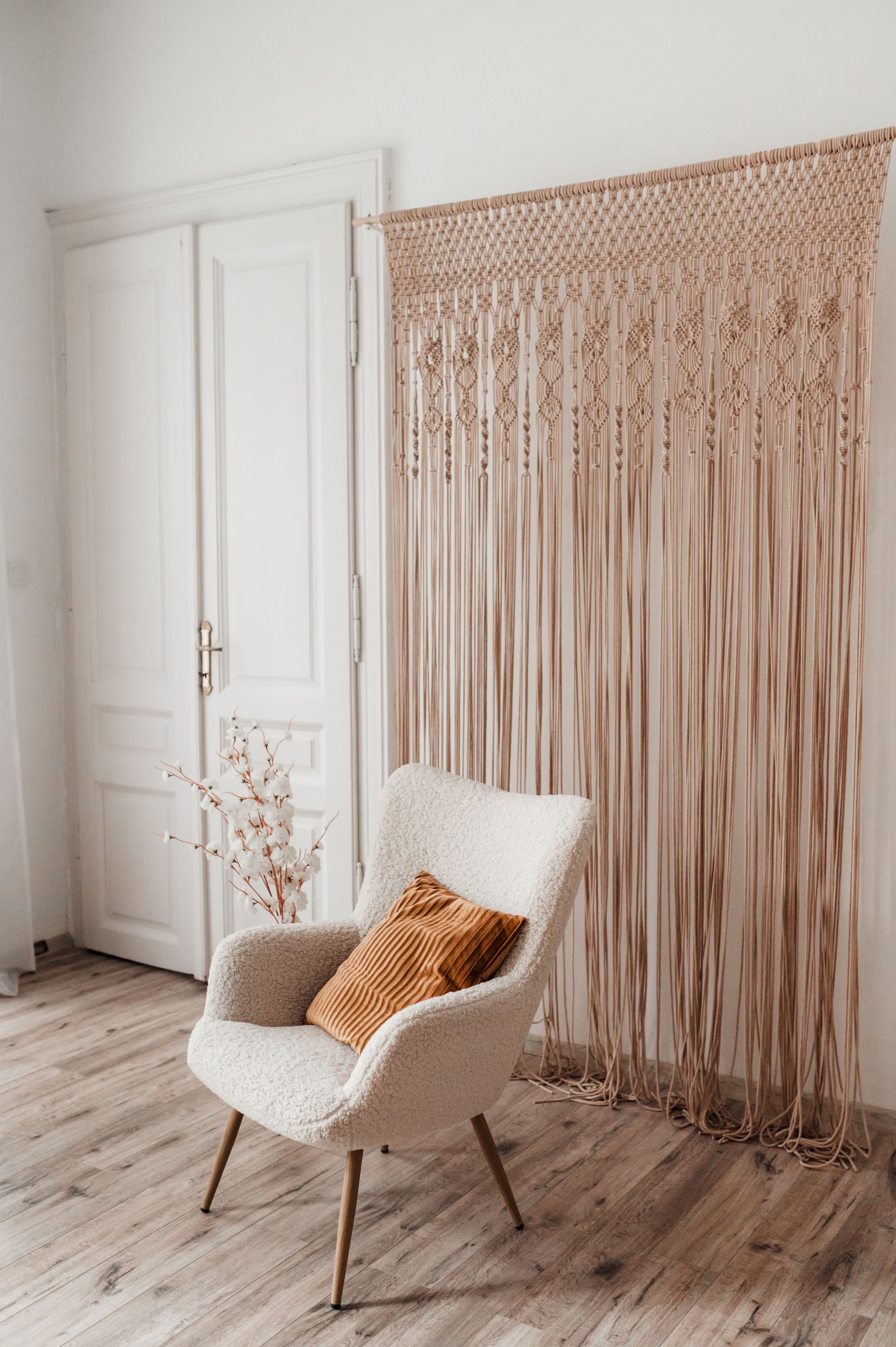A minimalist room design featuring a macrame wall hanging and cream accent chair with orange pillow.