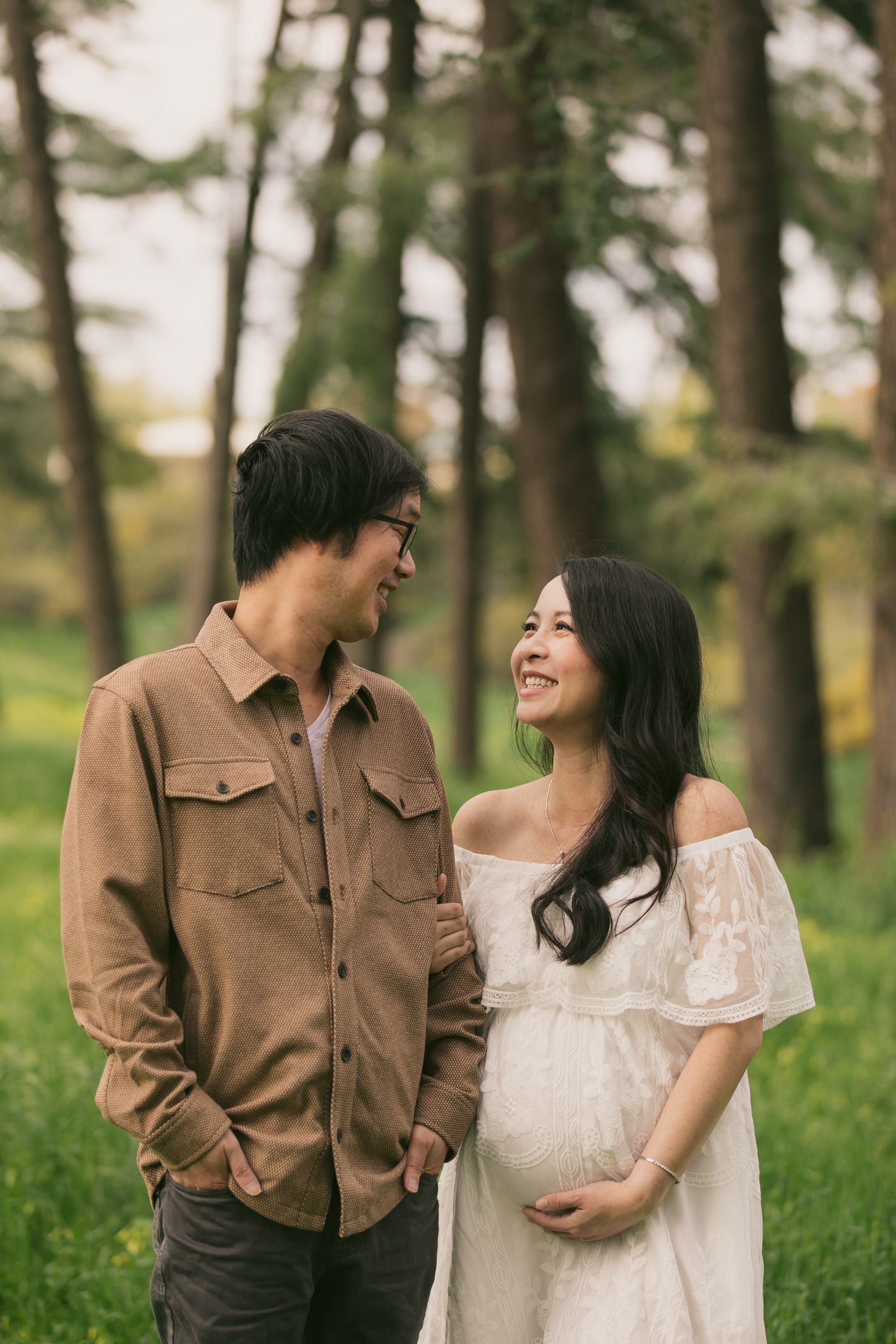 Expectant couple shares a tender moment in a wooded setting with the woman wearing a white lace dress.
