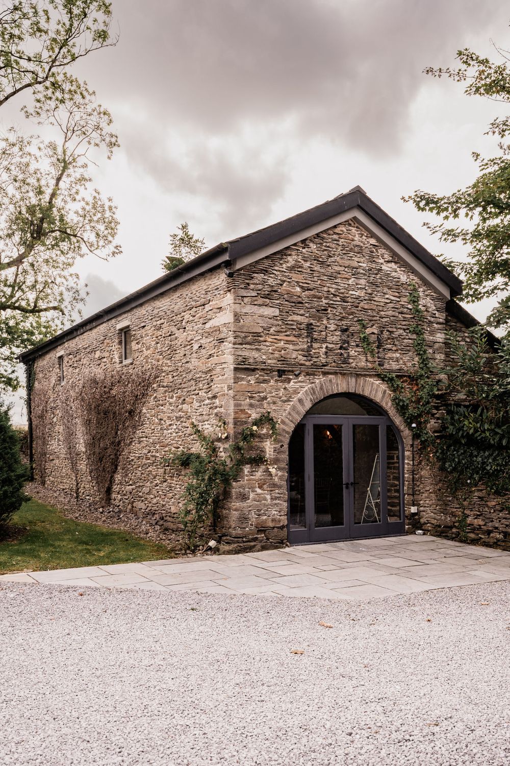 Rustic stone barn with arched entrance and gravel driveway surrounded by greenery.