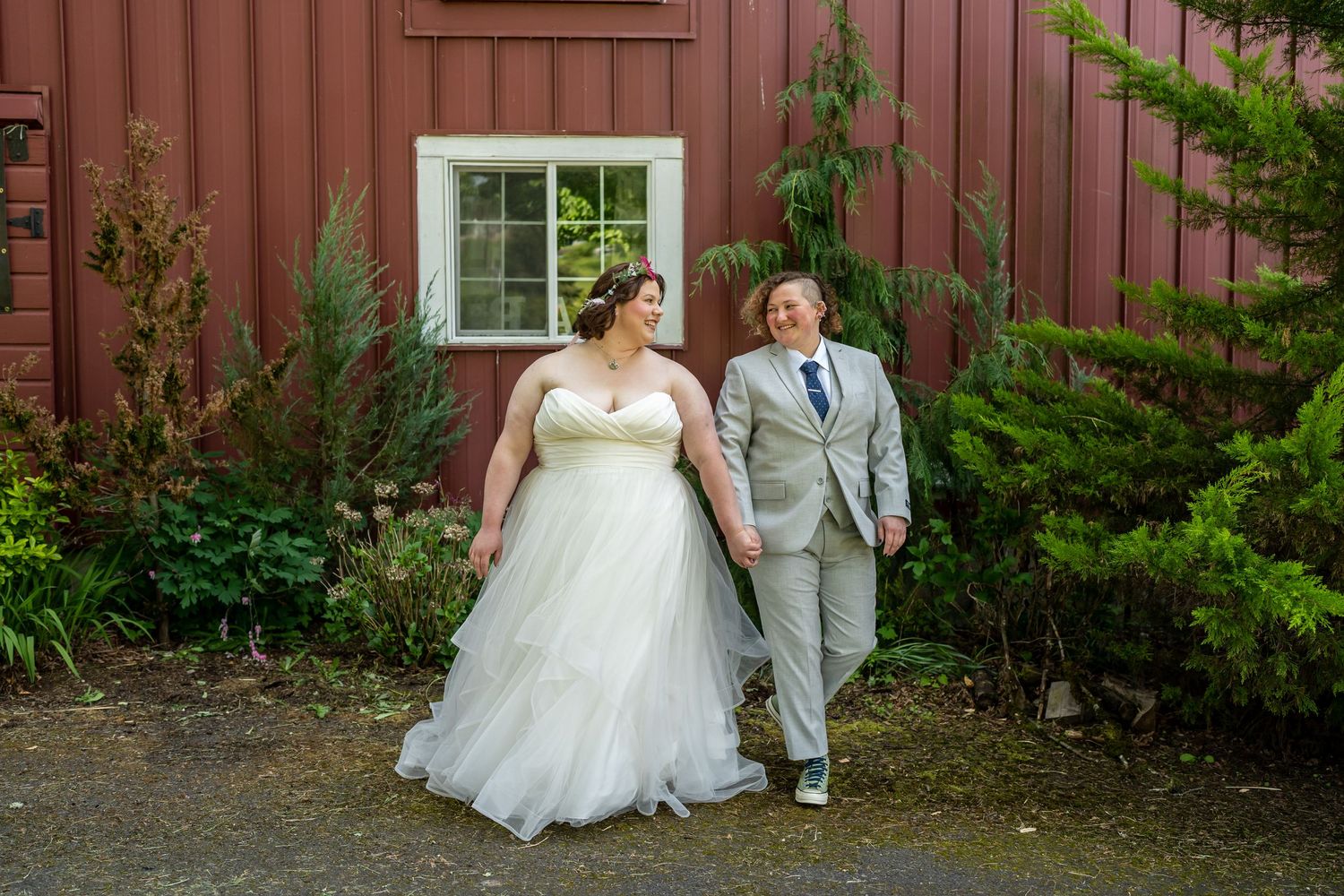 A couple smiles together in front of a rustic red barn with evergreen landscaping.