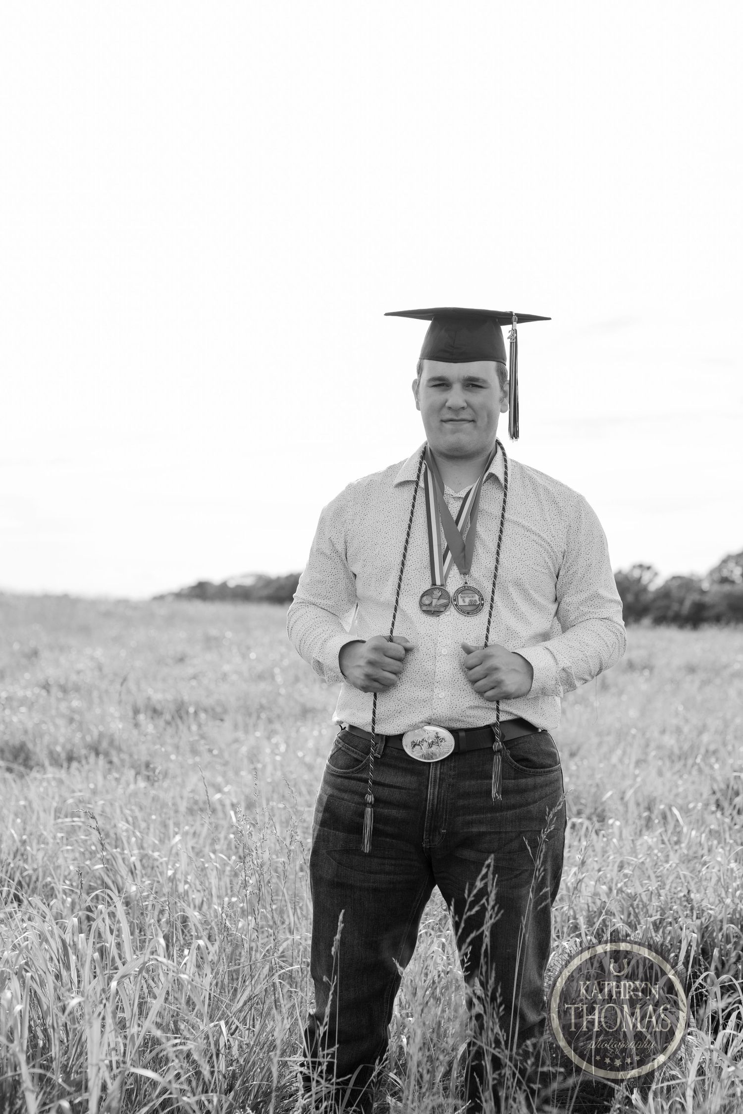 Black and white portrait of graduate in cap and gown standing in rural field.