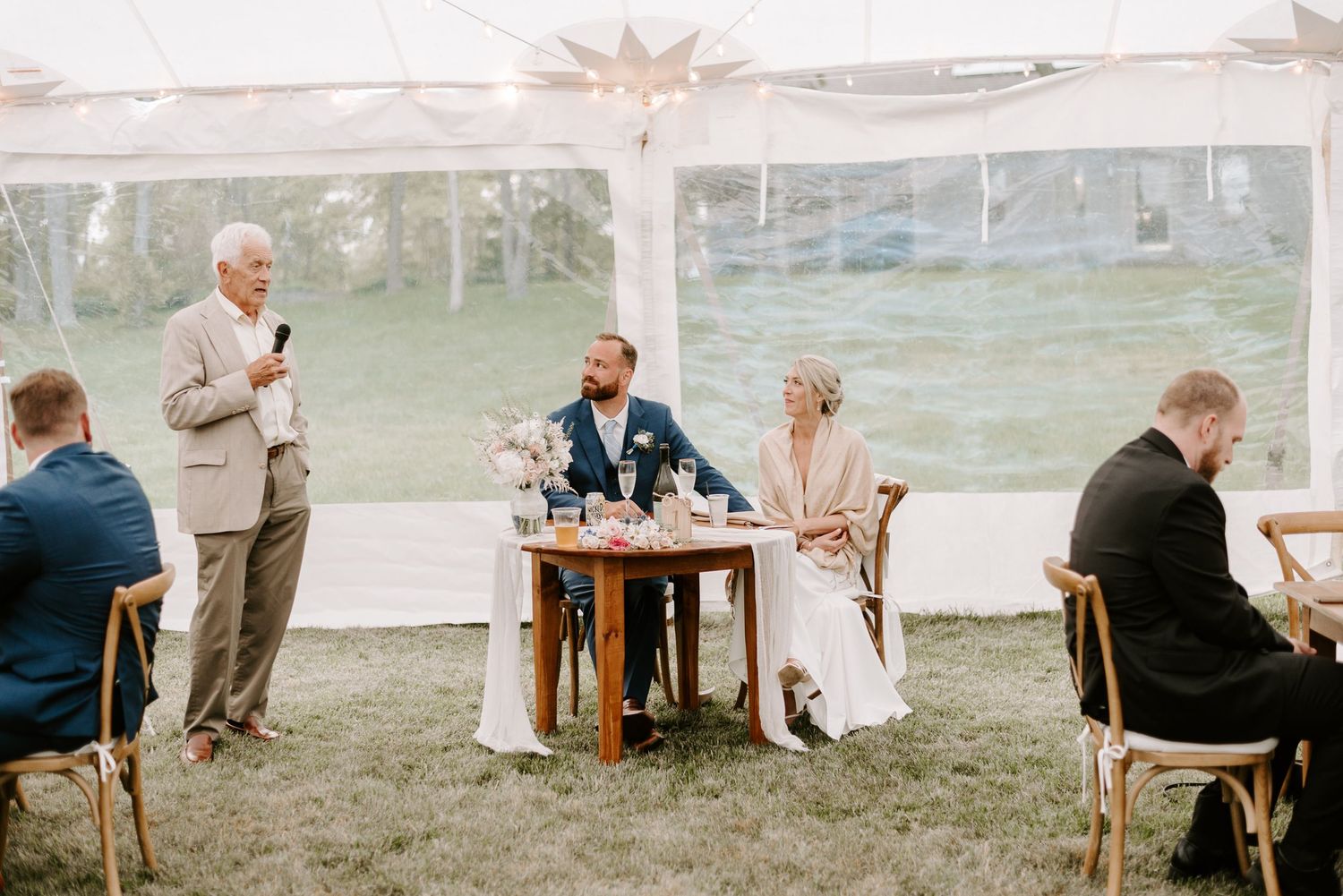 Outdoor wedding reception setup with rustic wooden tables under a white tent with string lights.