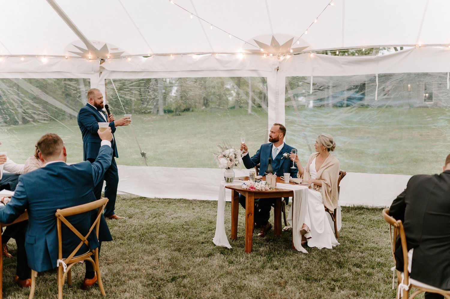 Wedding reception setup showing tables and chairs arranged under white tent on grassy lawn.