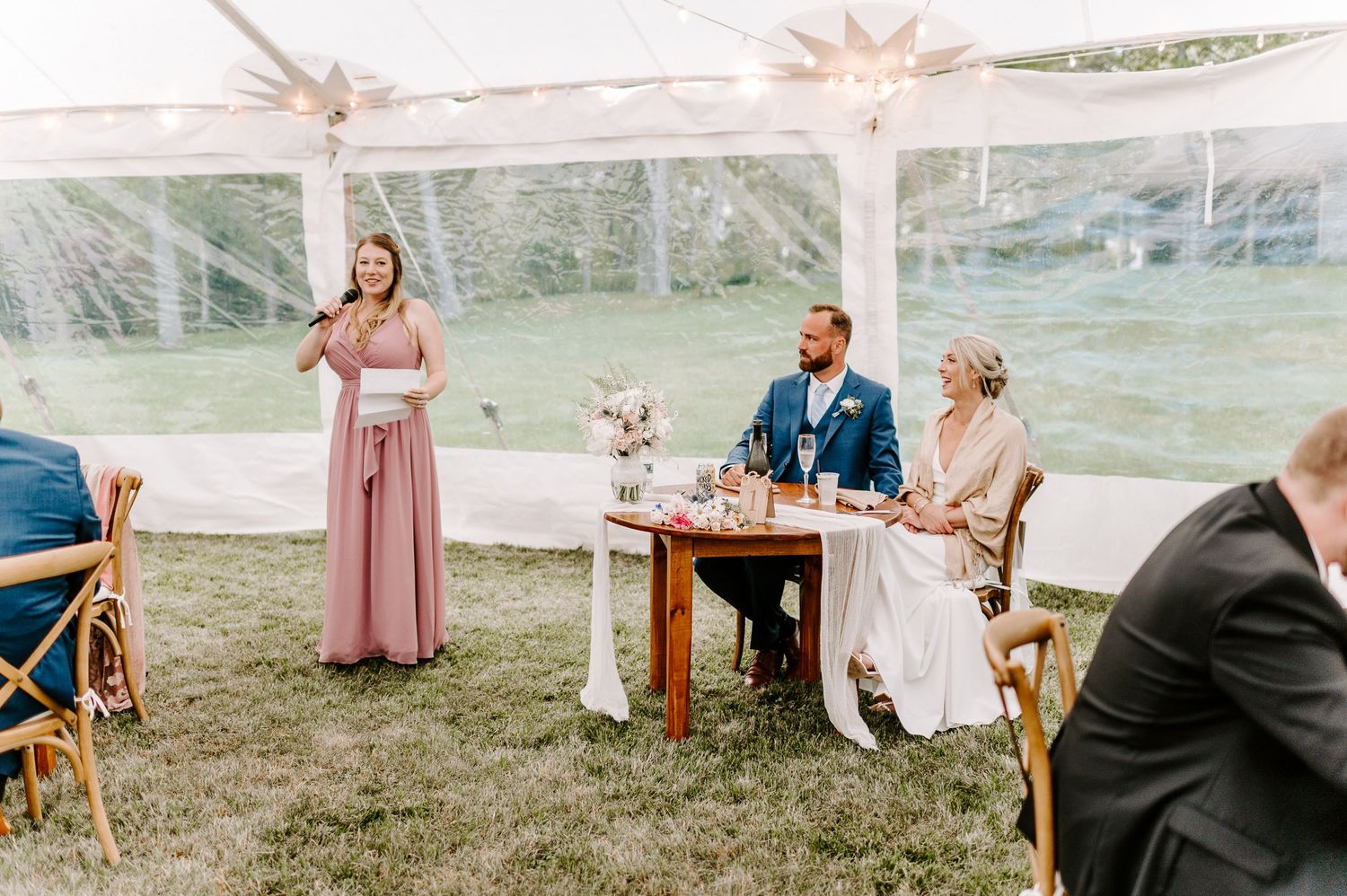 Elegant outdoor wedding reception with guests seated under white tent and string lighting.