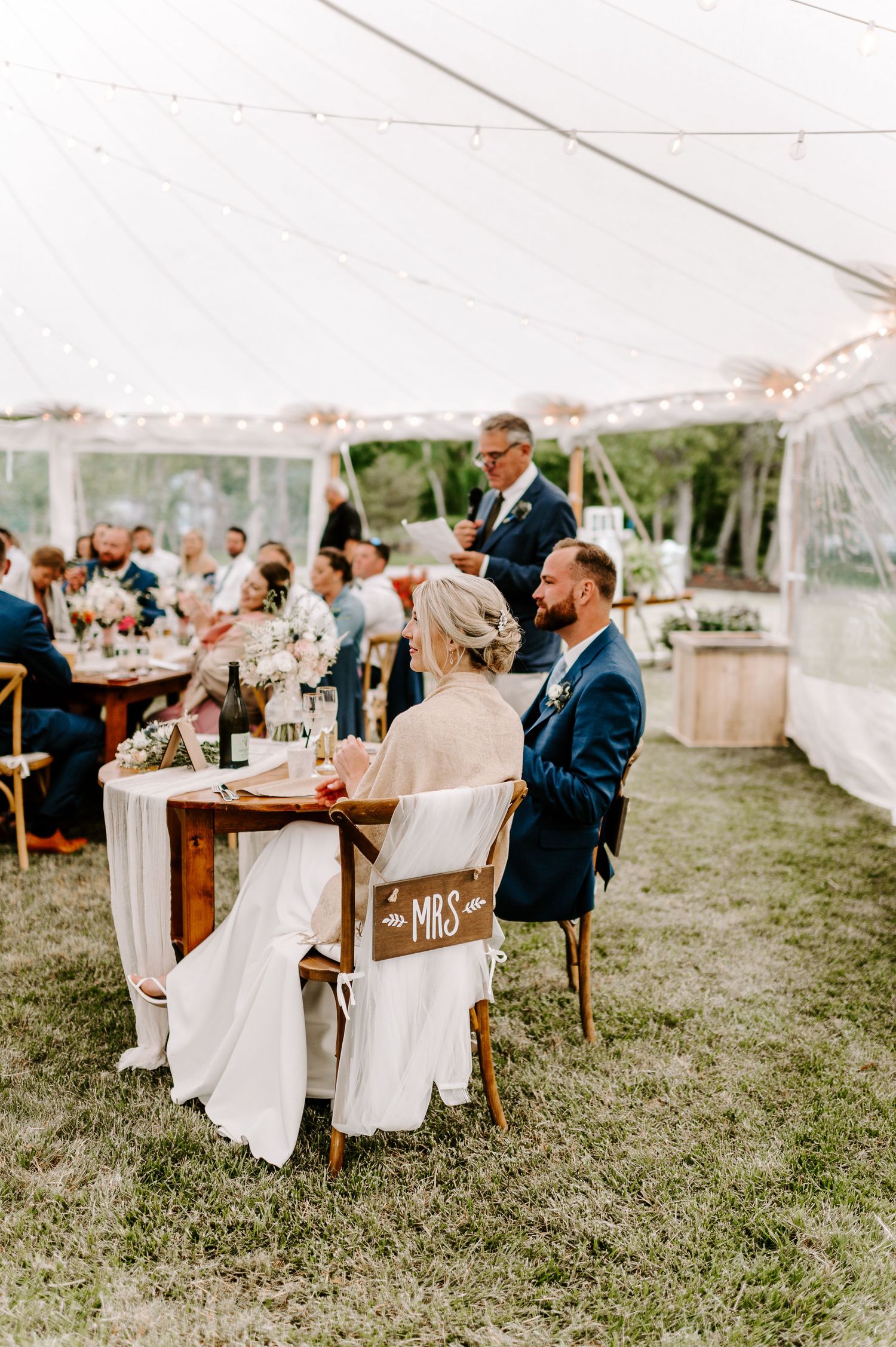 Wedding guests seated at wooden tables under tent with 'Mr and Mrs' chair signs visible.