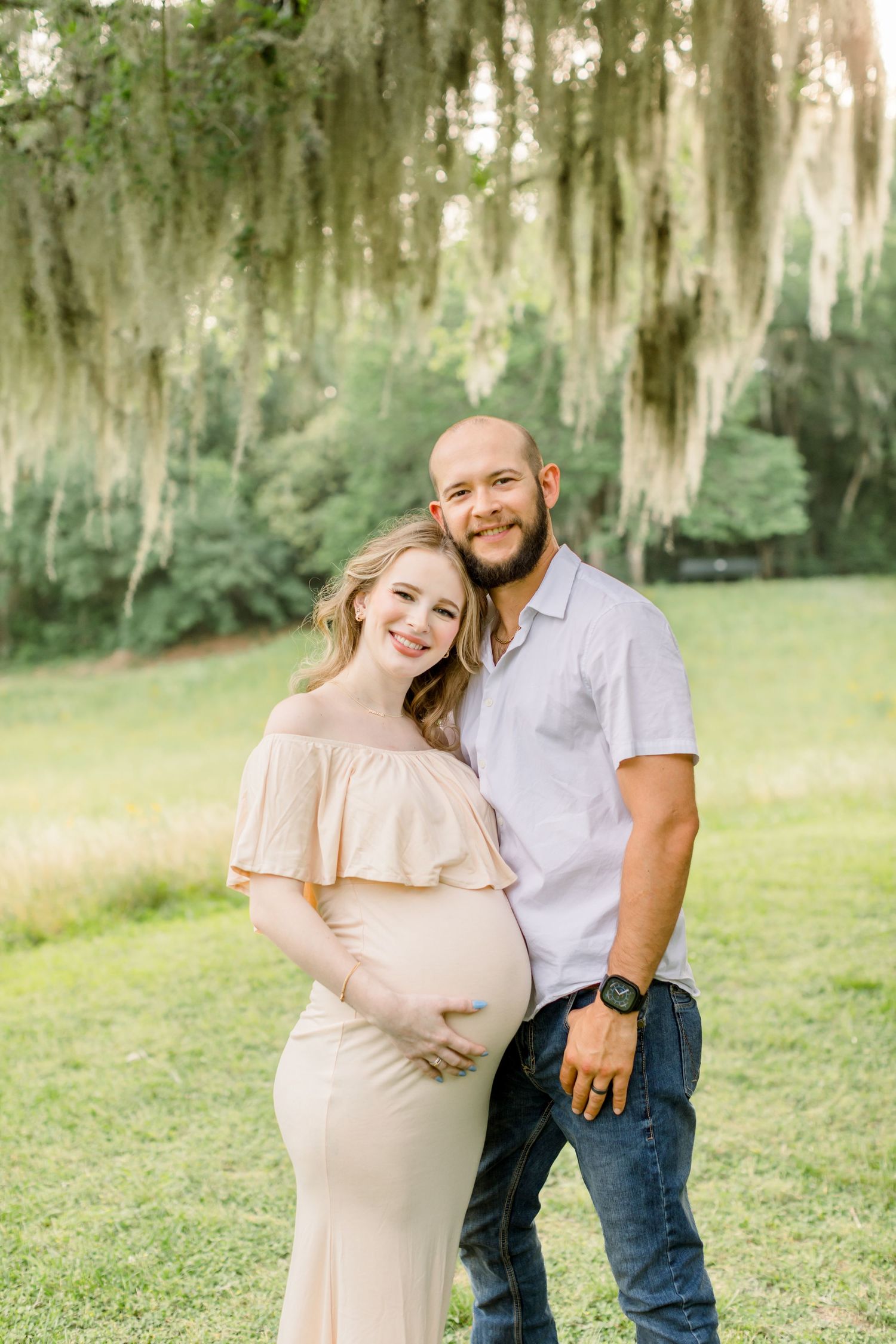 An outdoor maternity portrait under Spanish moss features a couple in casual attire.