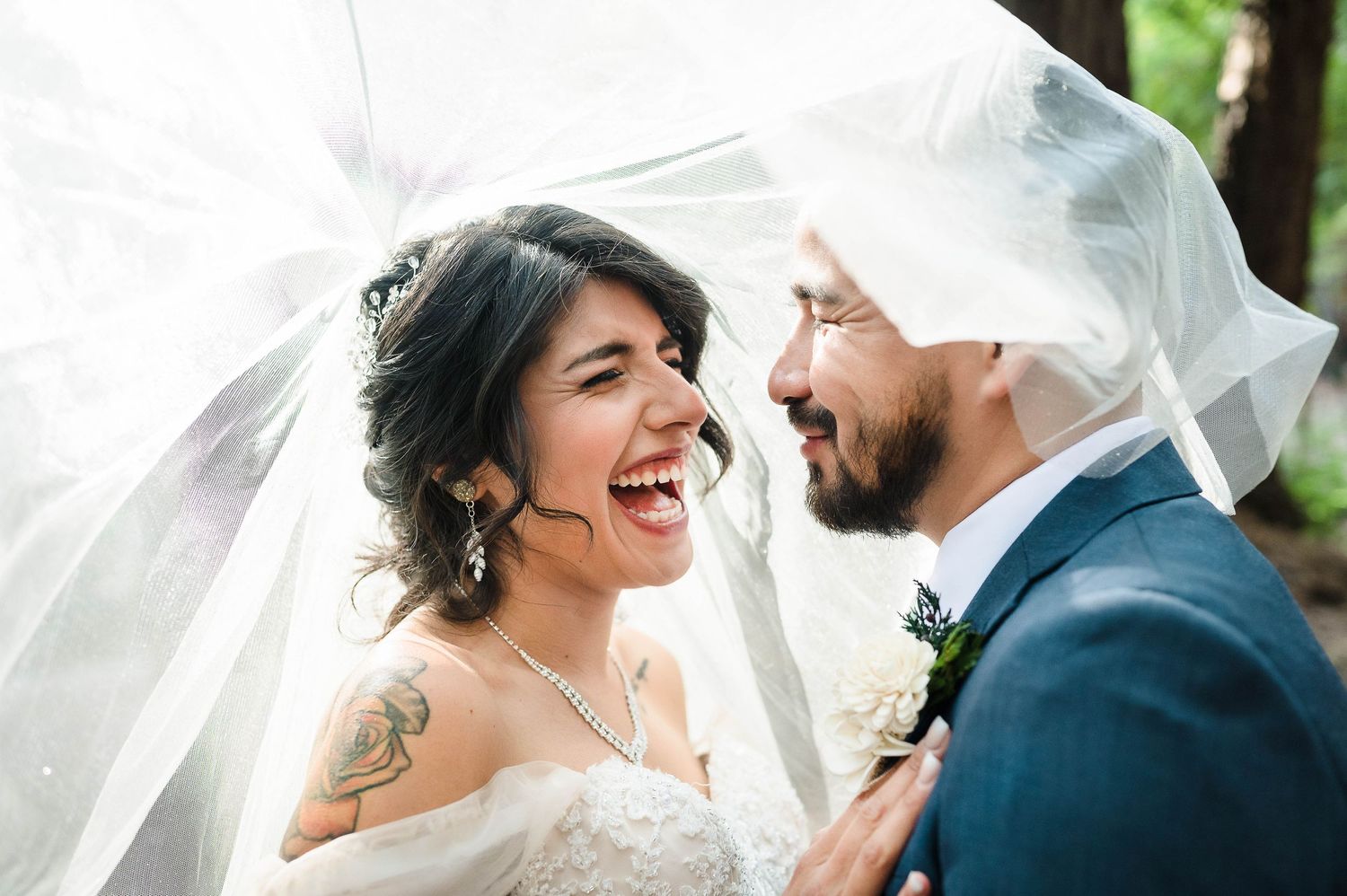 A couple laughs joyfully together under a flowing white veil during their wedding.