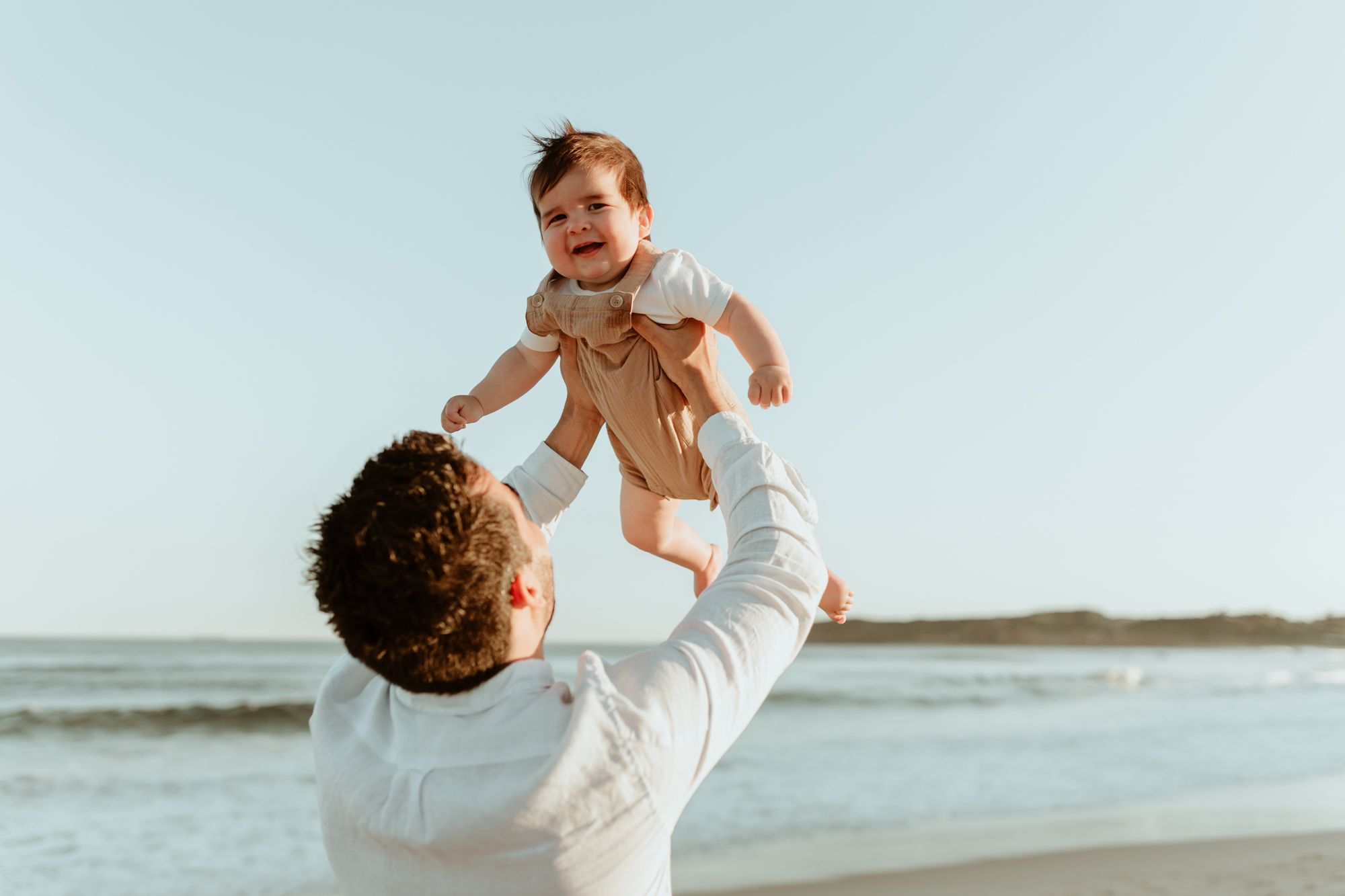 A joyful moment as a parent lifts their baby high into the air at the beach.