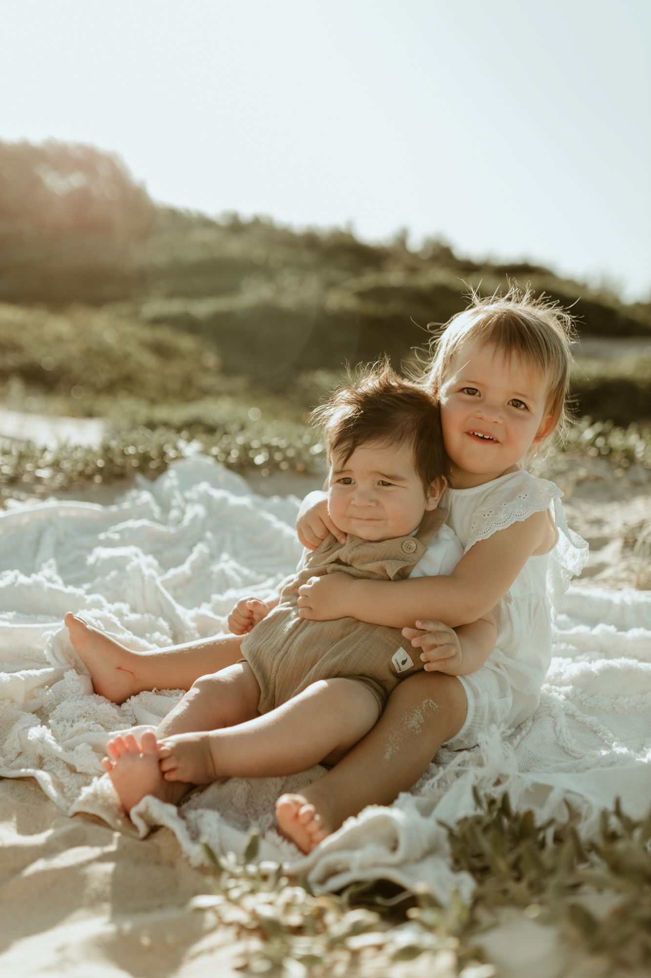 Two young children hugging and smiling while sitting on a white blanket at the beach.