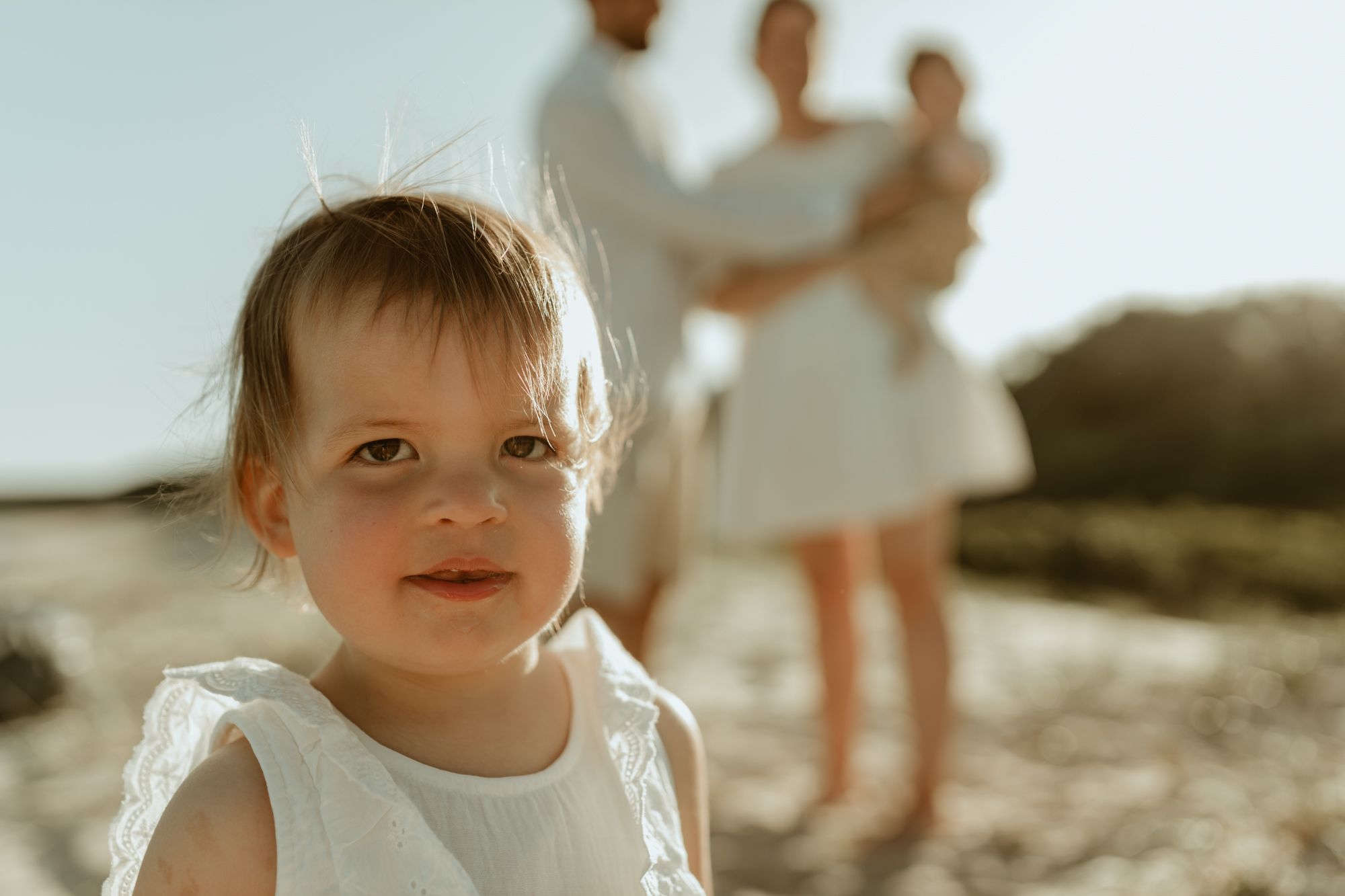 A toddler in white dress smiling at the beach with family in background.