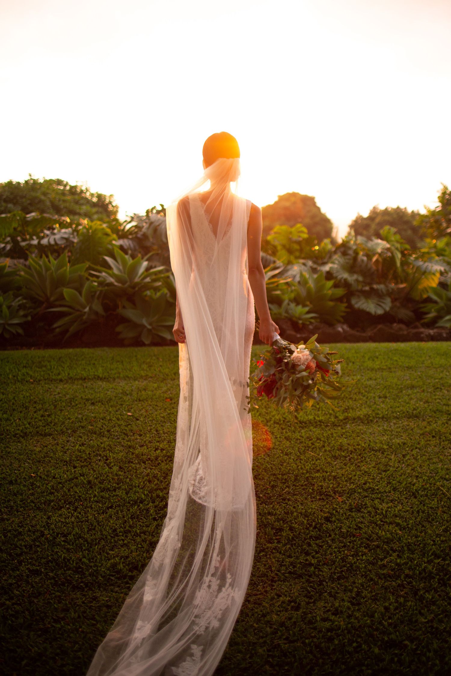 Silhouette of a bride in flowing dress against sunset in tropical garden.