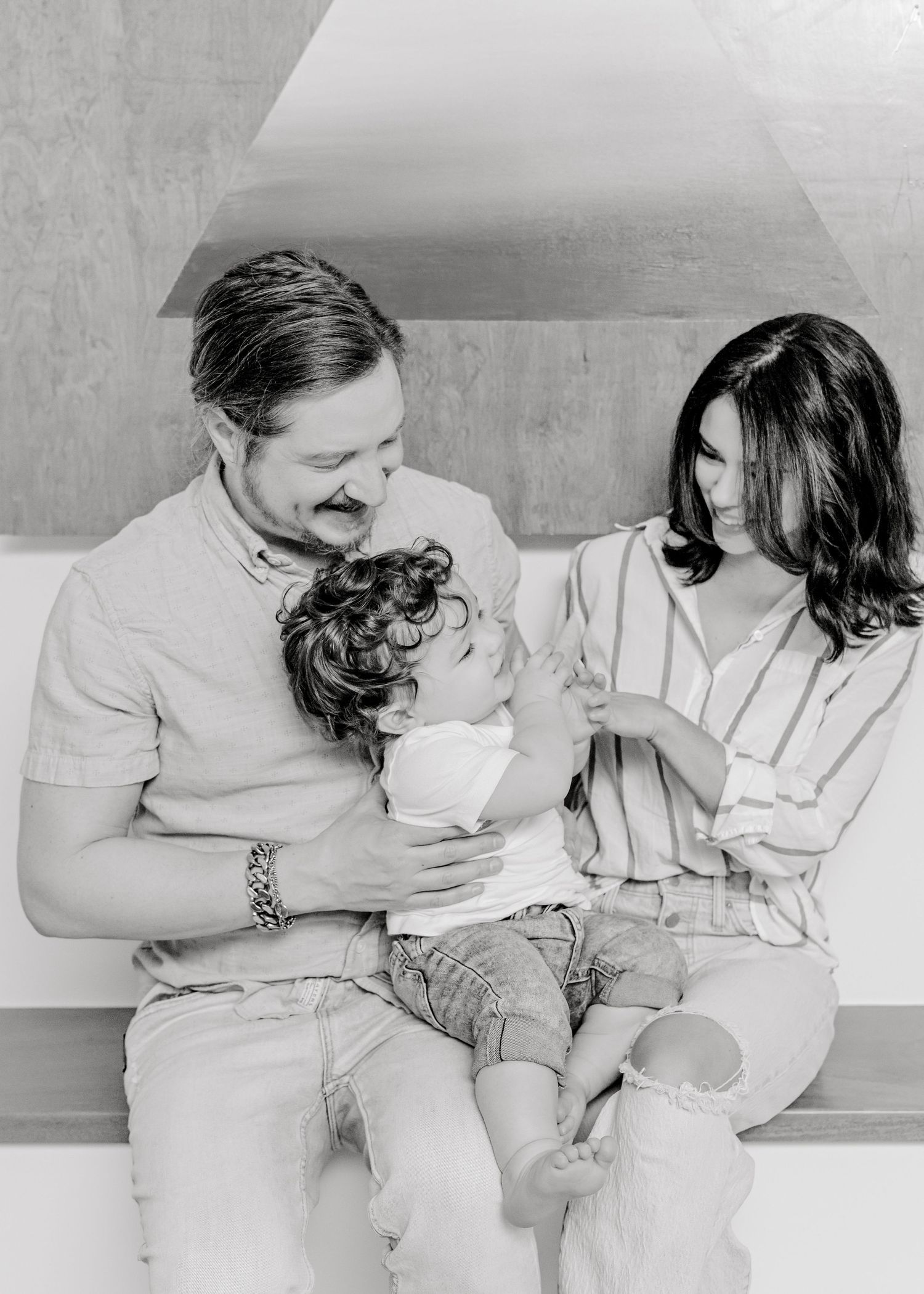 Austin, TX family sitting together on a bench in their home. dad holds their baby boy as mom tickles him