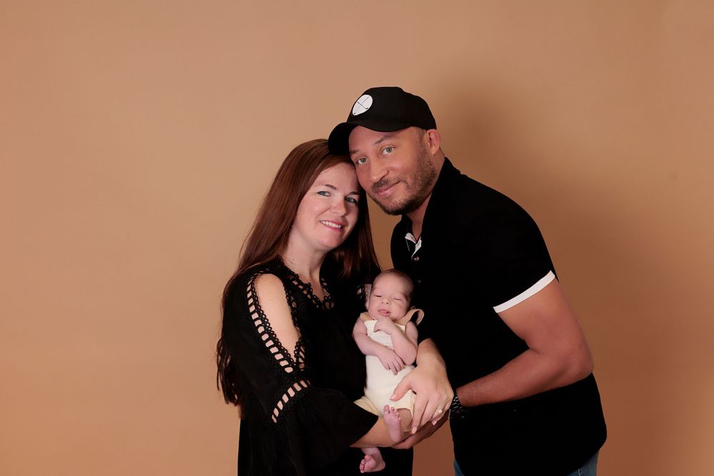 A couple poses for a studio portrait with their newborn baby against a brown backdrop.