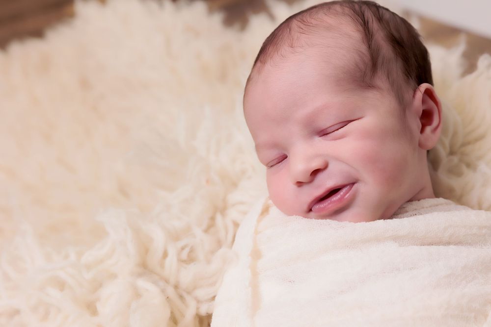 Smiling newborn baby wrapped in white swaddle on cream fur background.