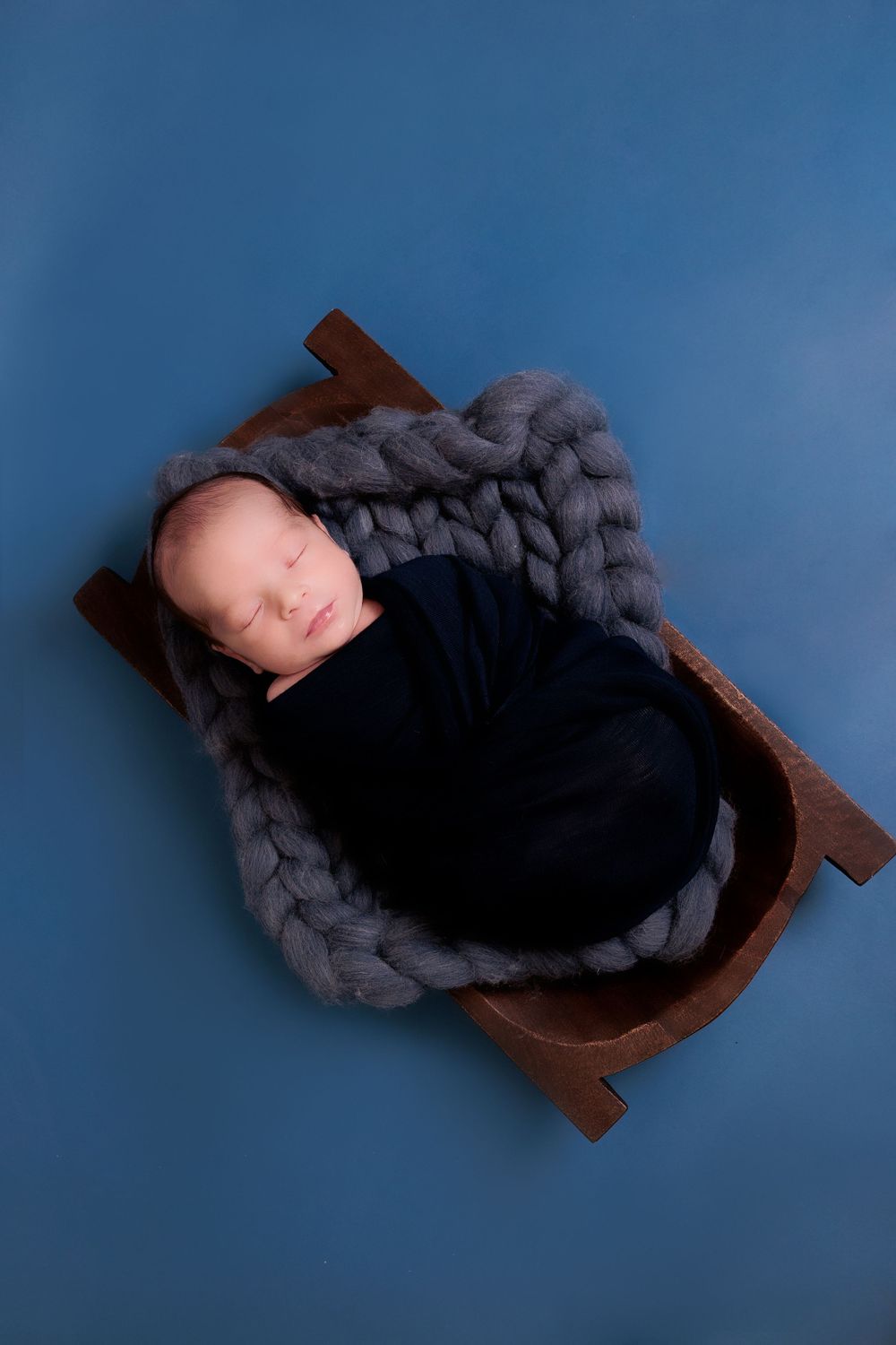 Peaceful newborn baby nestled in gray knit blanket on wooden prop against blue background.