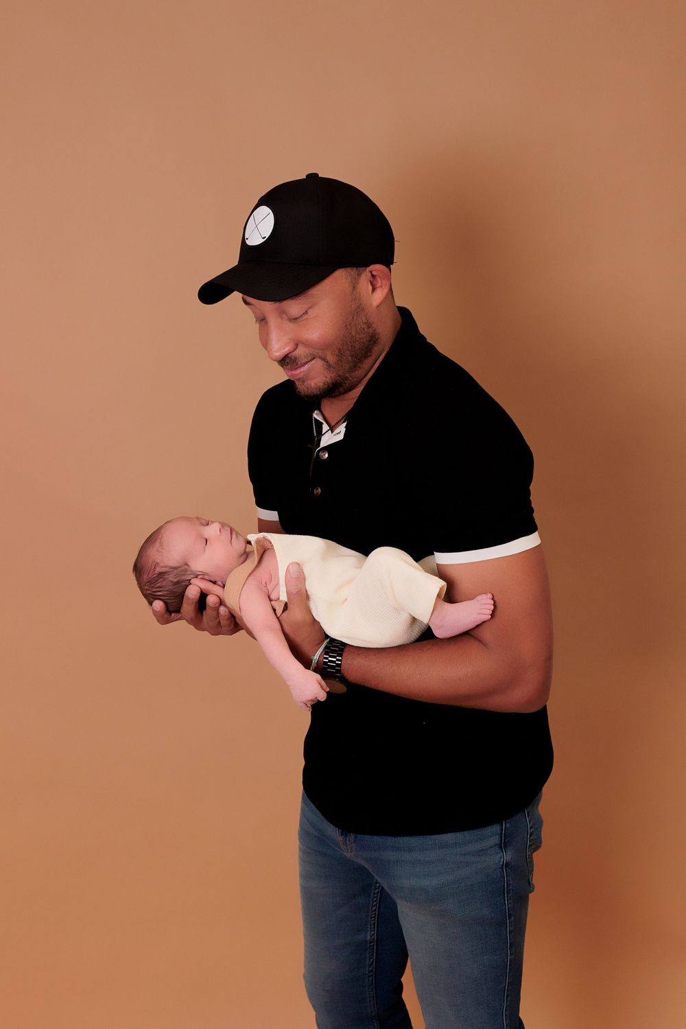 A person in a black shirt and baseball cap tenderly holds a sleeping newborn baby.