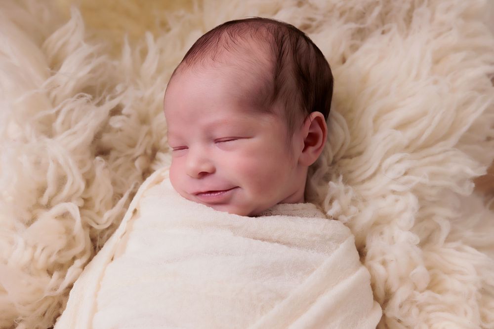 Peaceful sleeping newborn wrapped in white blanket on furry cream backdrop.