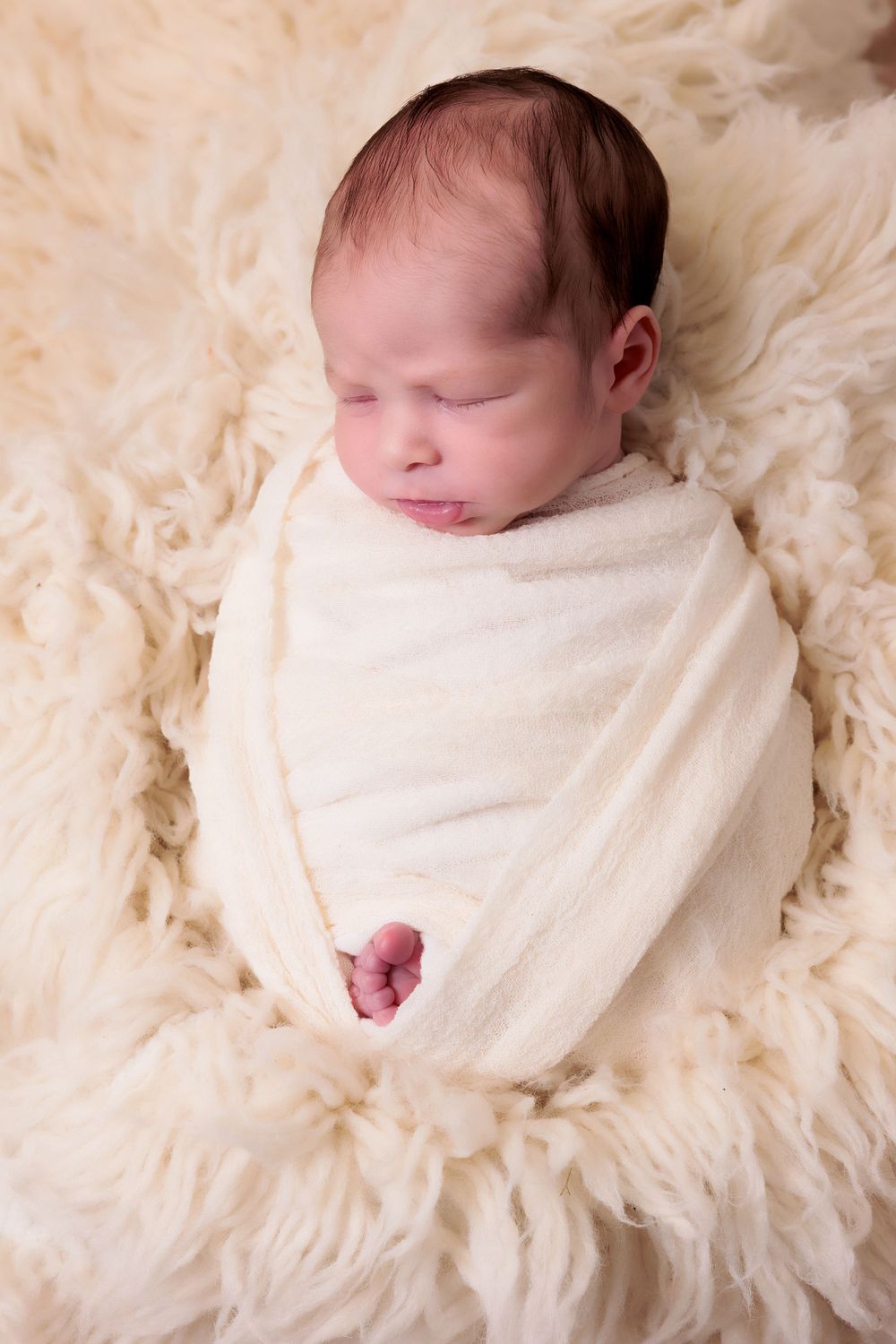 Sleeping newborn wrapped in white swaddle on fluffy cream fur backdrop.