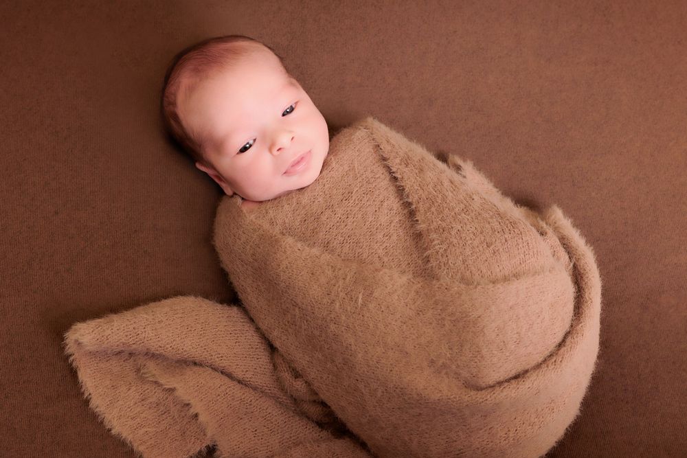 Sleeping newborn baby swaddled in soft brown blanket against brown background.