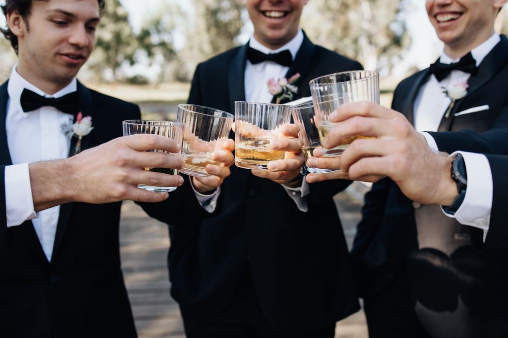 Groomsmen in black tuxedos and bow ties toasting with glasses during wedding celebration.