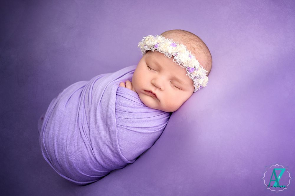 Peaceful newborn portrait with lavender wrap and floral crown against purple background.
