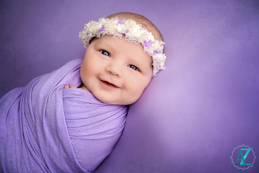 Joyful newborn portrait with purple wrap and floral headband against matching background.