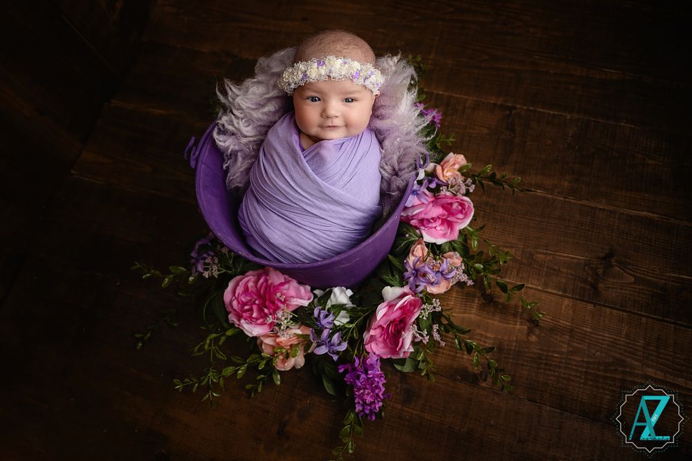 Overhead newborn photo with purple wrap and pink floral decorations on wooden floor.