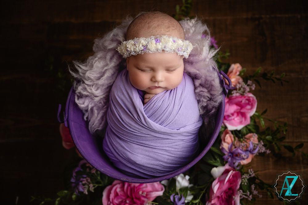 Artistic newborn photo in purple bowl with floral decorations on wooden background.
