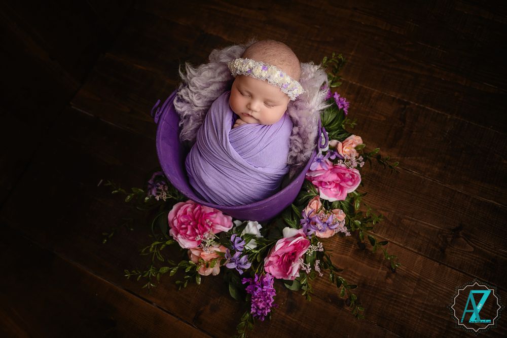 Newborn swaddled in purple fabric rests in a bowl surrounded by pink and purple flowers on wooden floor.