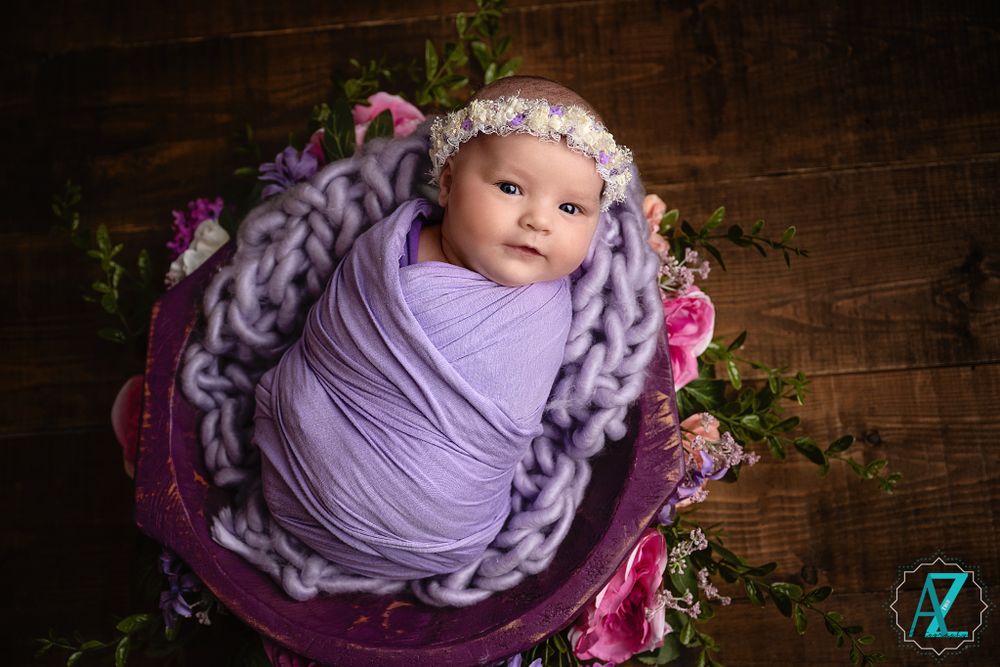 Sleeping infant wrapped in lilac fabric nestled in a purple bowl with floral decor.