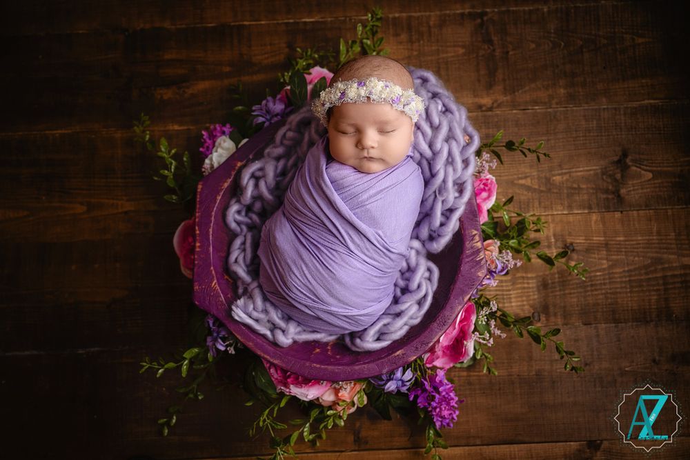 Newborn in purple wrap sleeps in bowl with chunky knit blanket and pink florals.