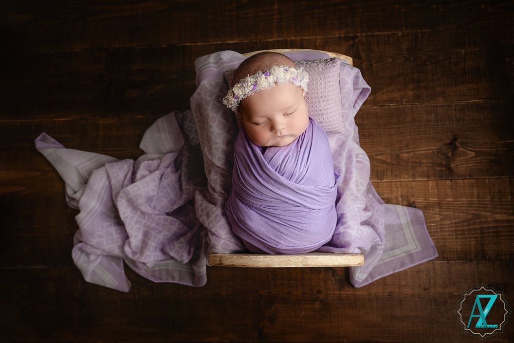 Infant swaddled in lavender fabric rests on wooden platform with soft textiles.