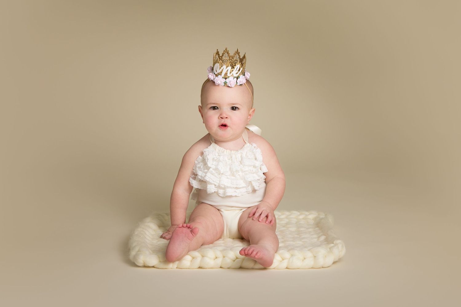 Baby wearing crown and cream dress sitting on neutral backdrop for portrait session.