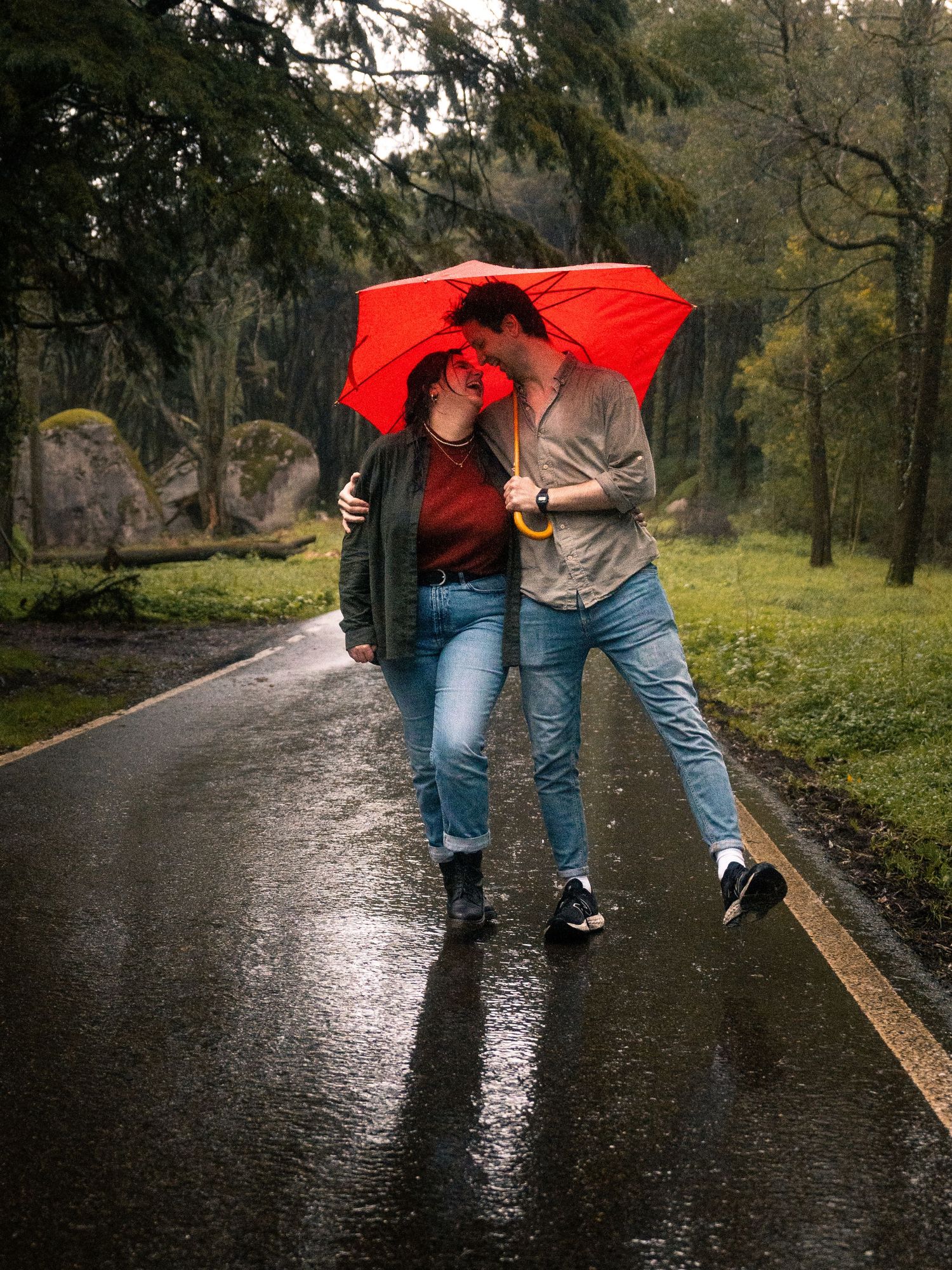 Couple sharing a red umbrella on a rainy path through a wooded park.