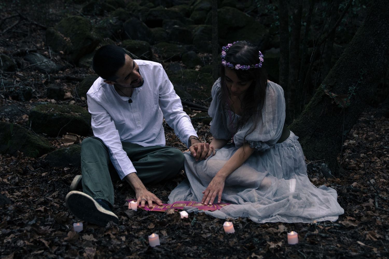 Candlelit romantic scene in a dark forest setting with couple in white and grey attire.