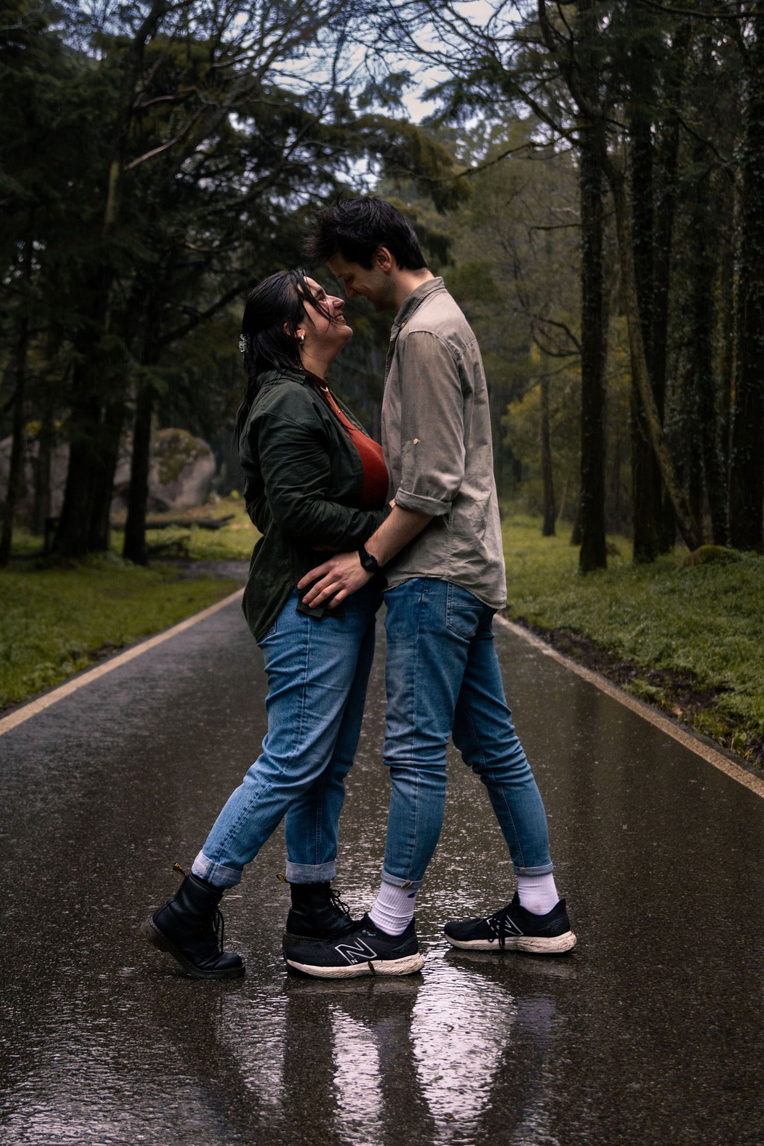 Couple in jeans dancing on wet pavement in wooded area.