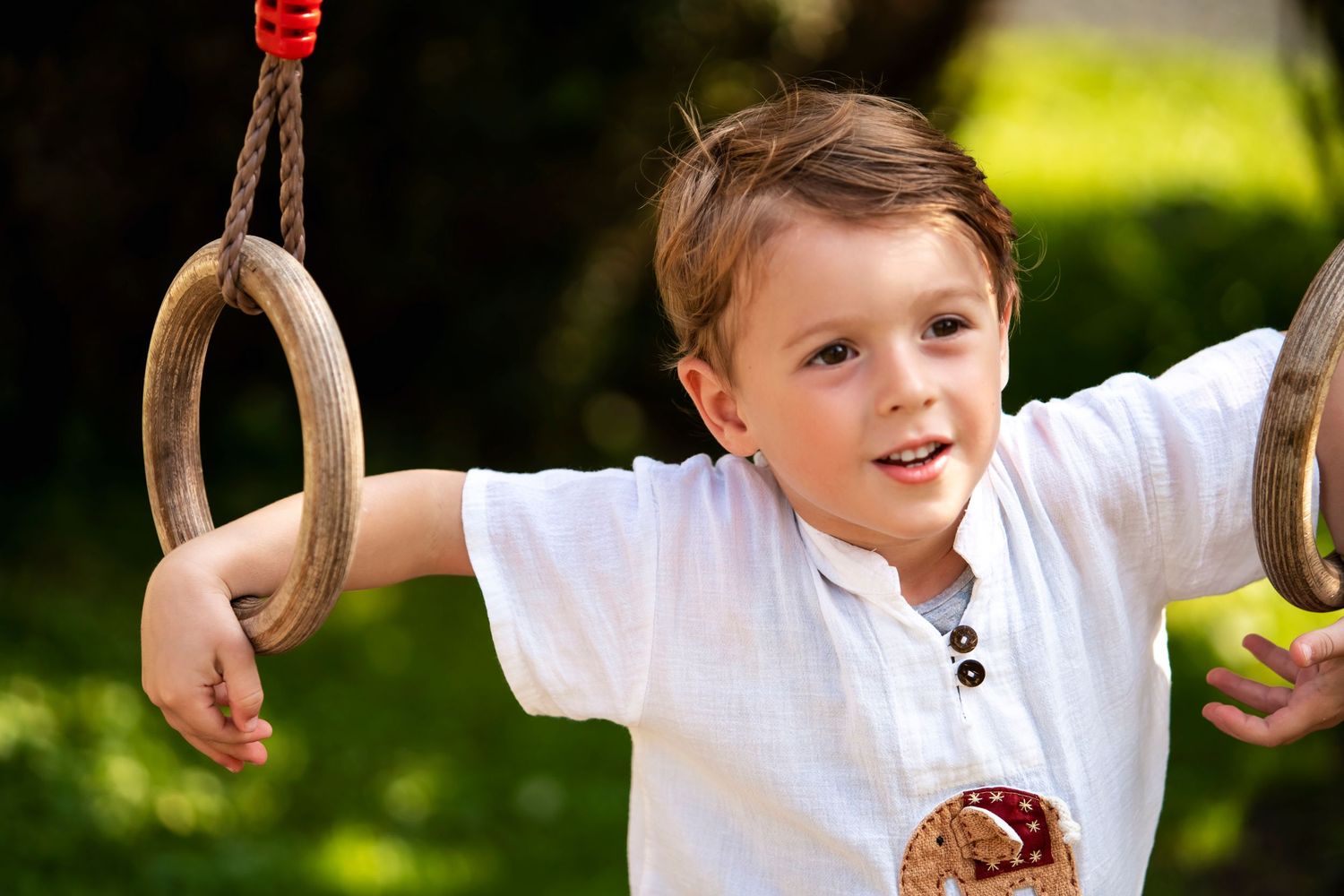 A cheerful child plays on wooden gymnastic rings in a sunny garden.