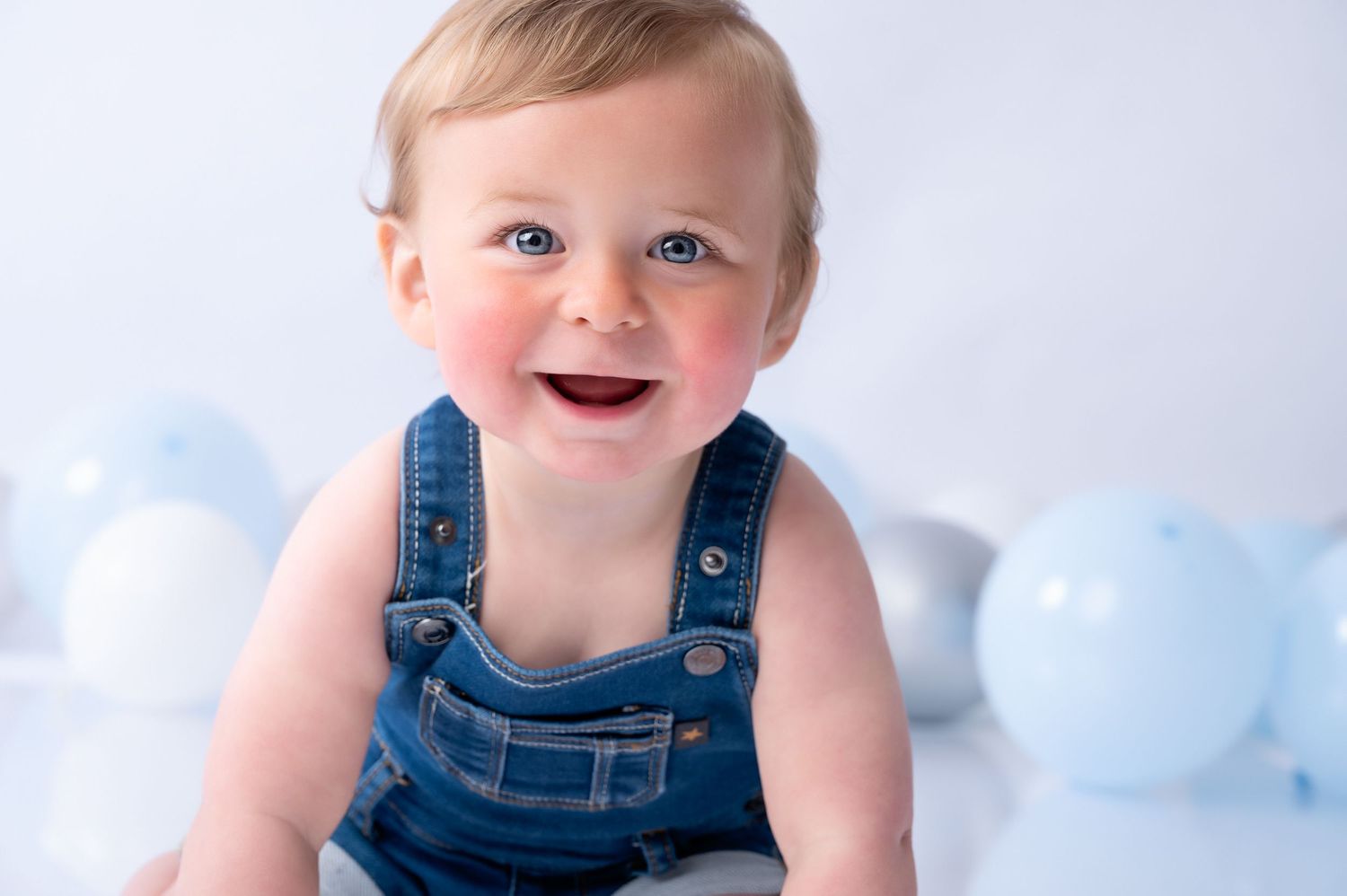 Smiling baby in denim overalls sitting against blue balloons background.
