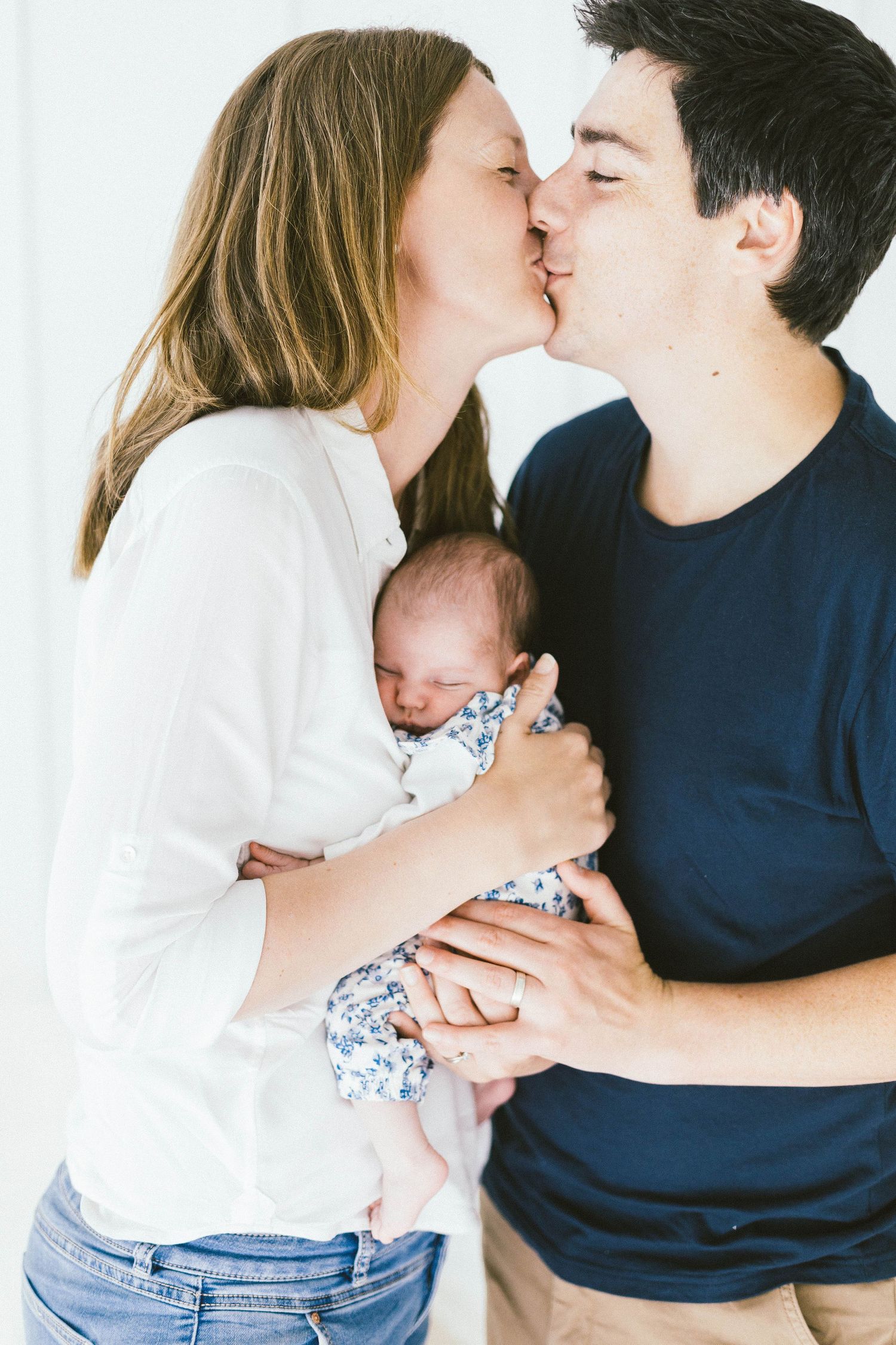 Parents share a kiss while cradling their sleeping newborn baby in a floral outfit.