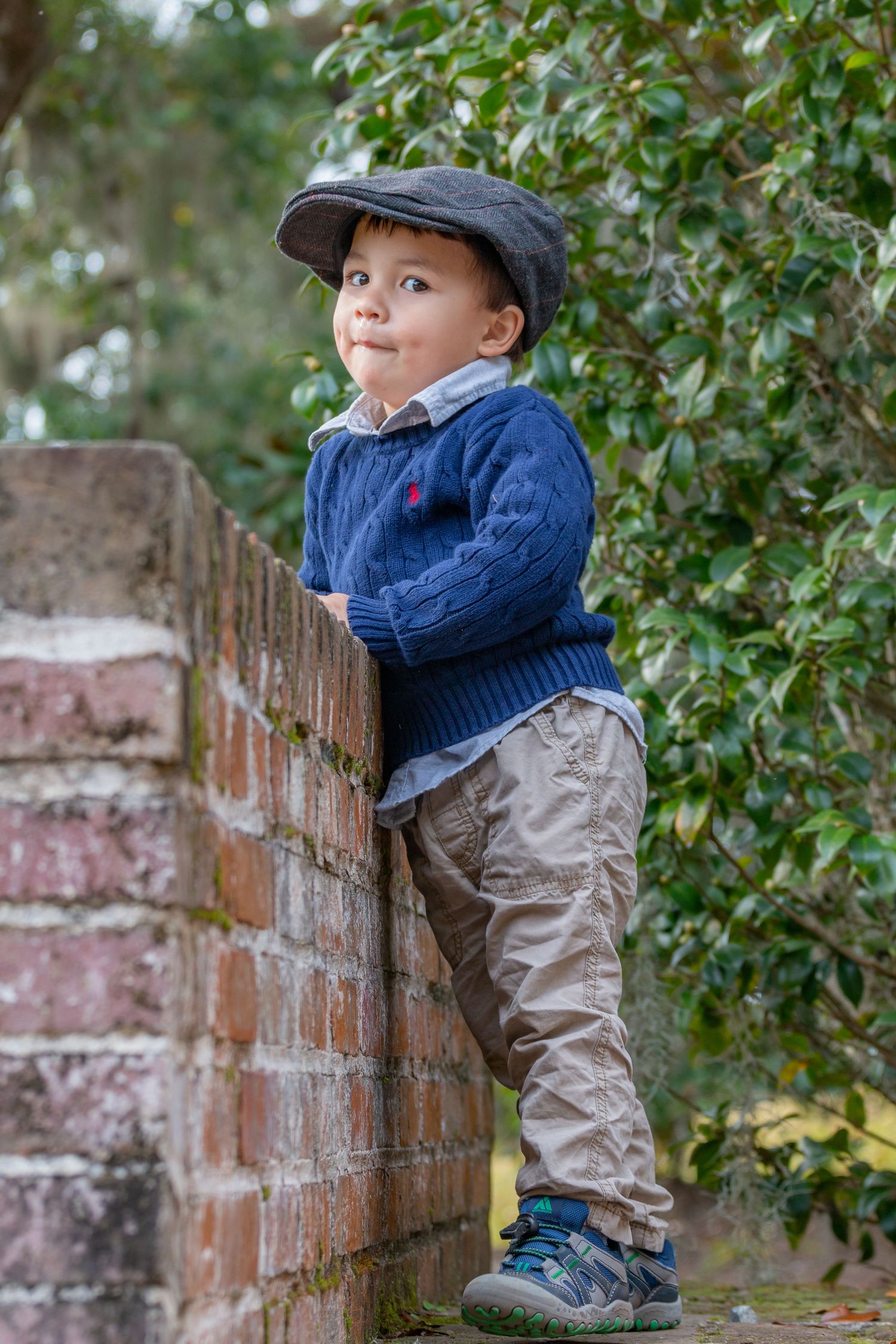 A small child wearing a cap and sweater poses playfully on a brick wall outdoors.