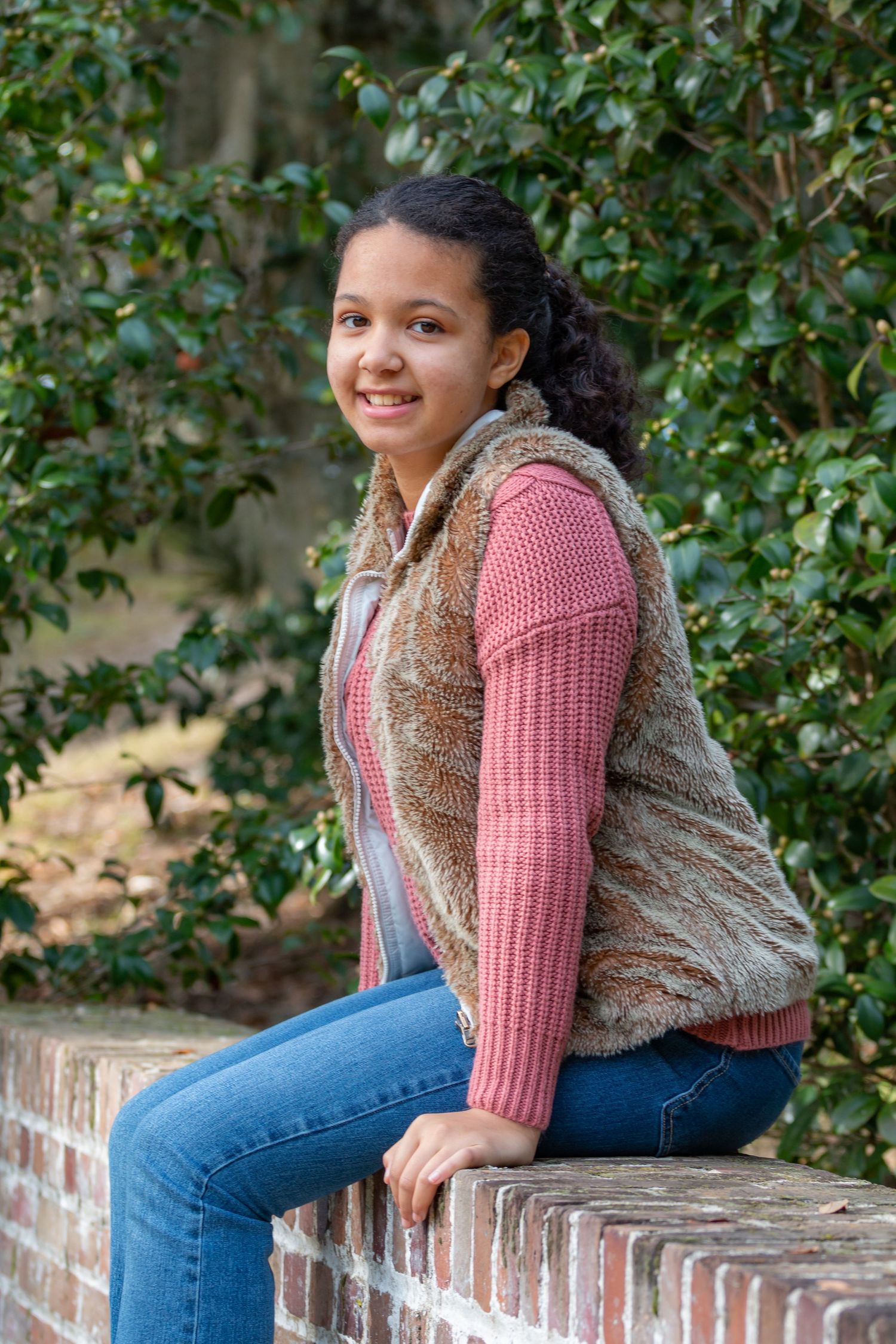 A person in a pink sweater and fur vest sits casually on a brick wall surrounded by greenery.