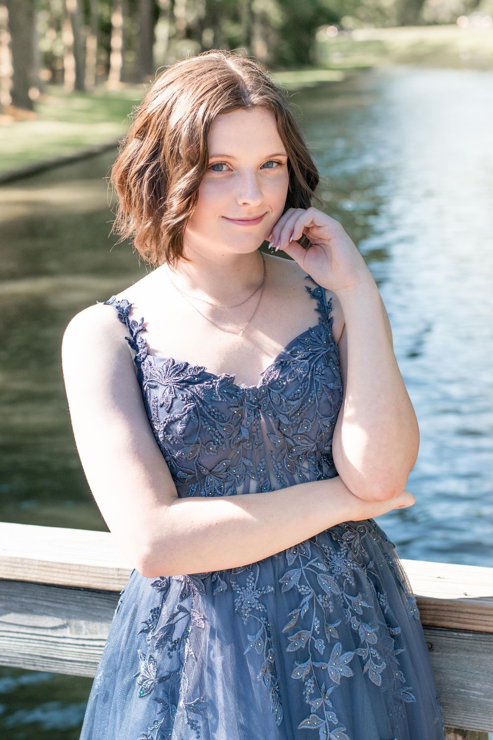 A elegant blue lace formal dress photographed by a river with trees and sunlight in the background.