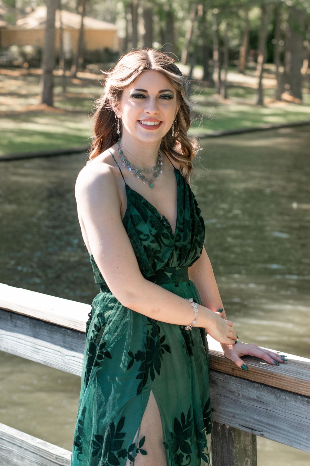 Woman in emerald green formal dress poses on wooden bridge railing.