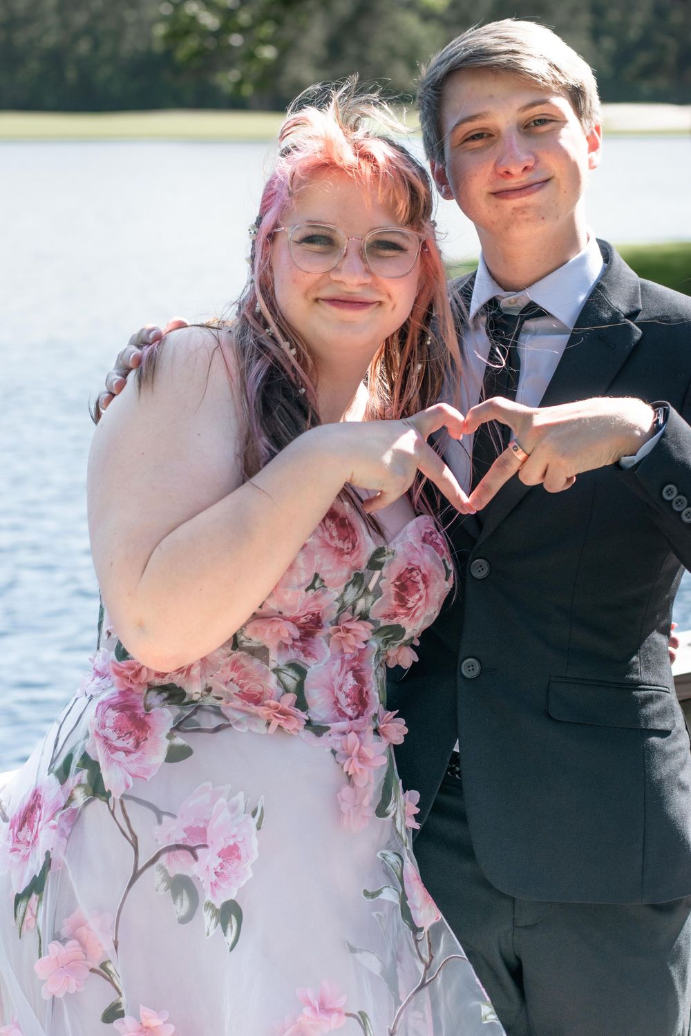 Couple in formal wear making heart shape with hands by lakeside.