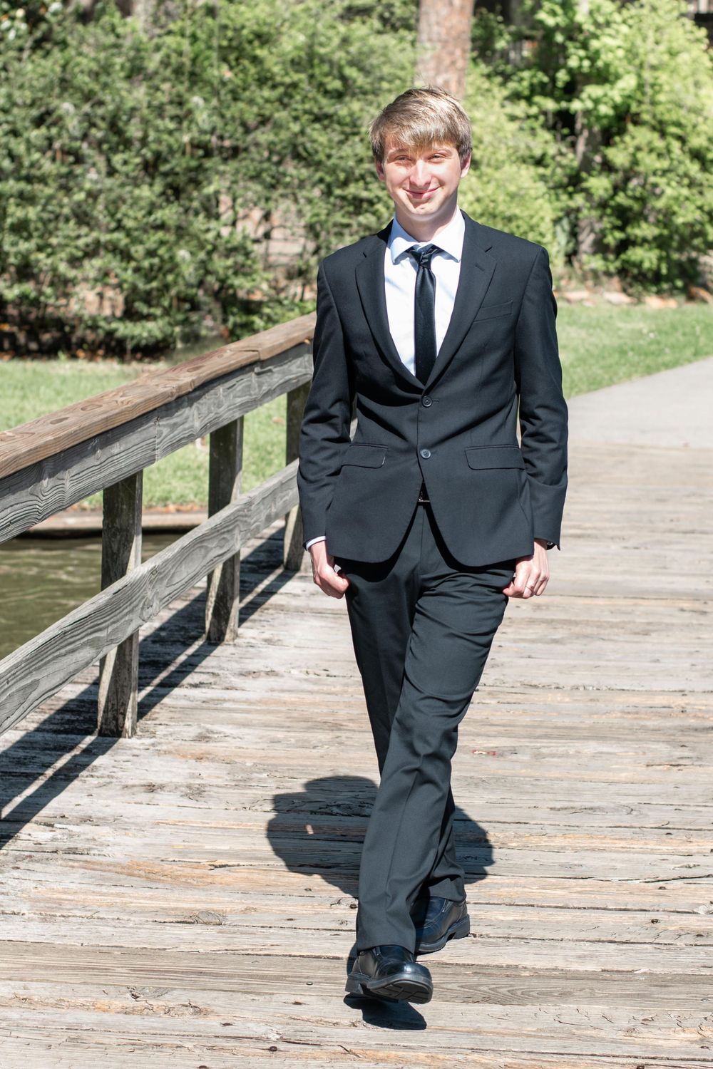 Person in black suit poses on wooden boardwalk in sunny outdoor setting.