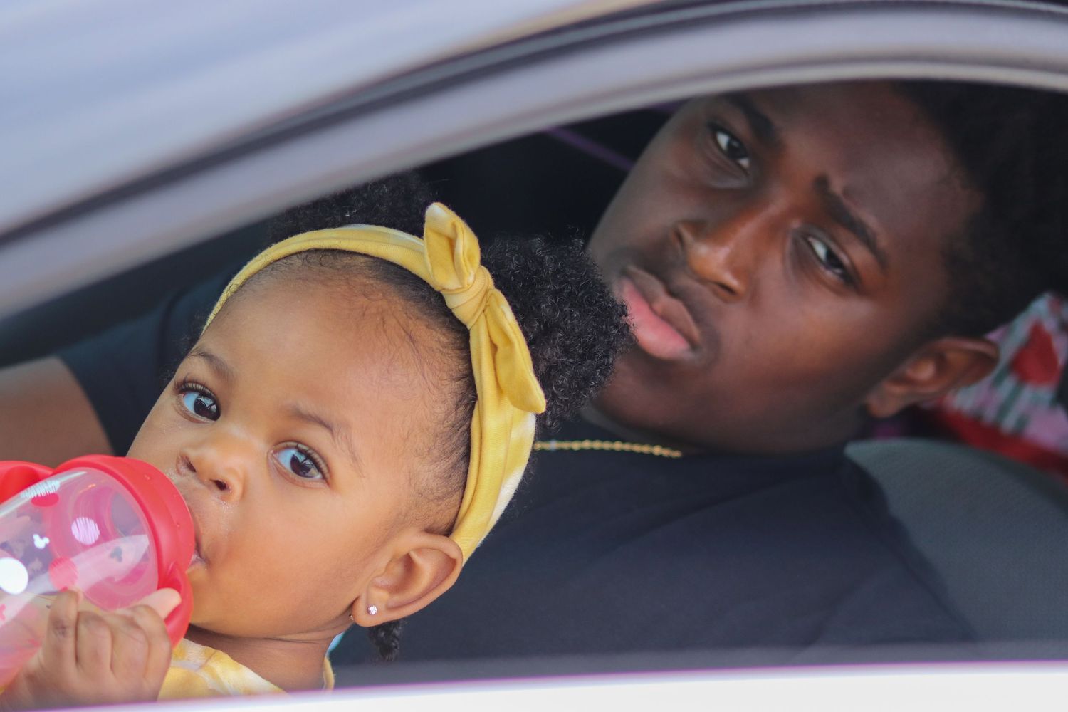 Baby with yellow headband drinking from bottle while sitting in car with adult.