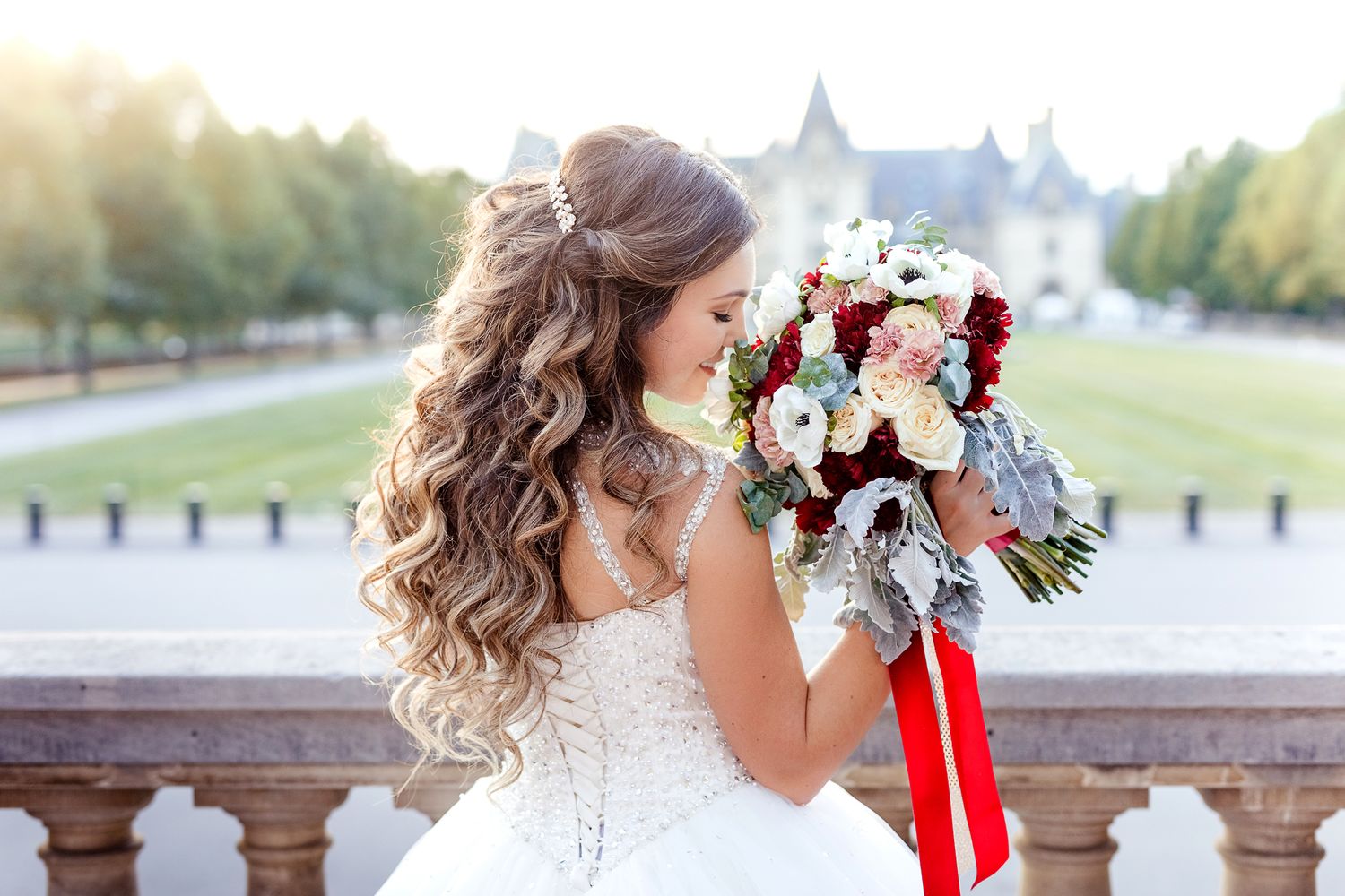 Bride with flowing curls holding a red and white bouquet against a scenic background.