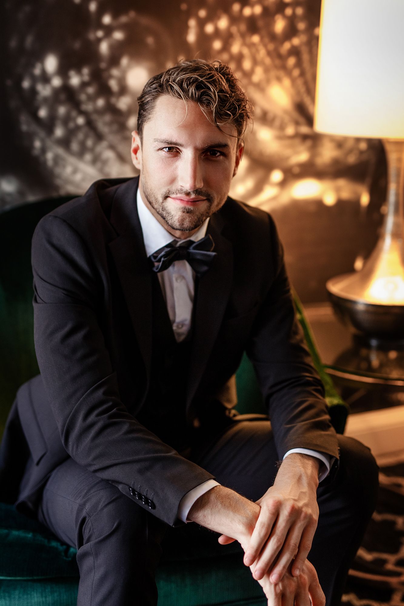 Sophisticated formal portrait of gentleman in classic black tuxedo seated on emerald velvet chair.